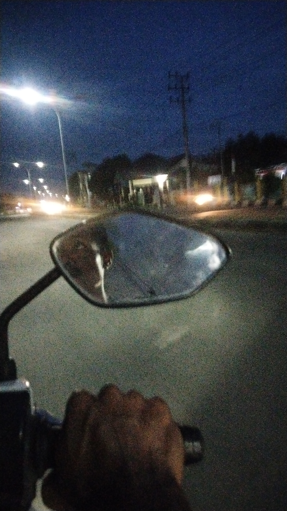 A first-person perspective shot from a motorcycle in Lhokseumawe, Indonesia, captures a nighttime scene on a paved road. The sky is a dark, grainy blue, indicative of a photograph taken in low light conditions. Streetlights cast a bright glow on the left side of the image, illuminating the road ahead. In the distance, the headlights of other vehicles create streaks of light, suggesting traffic. Buildings are visible along the right side of the road, with some lights illuminated within. A utility pole with crossbars stands prominently against the sky on the right.
In the foreground, a hand is visible, gripping the handlebar of the motorcycle. The motorcycle's side mirror is in focus, reflecting a fractured image of the road and possibly the rider. The mirror itself appears to be damaged, with visible cracks. The overall image has a grainy texture, typical of nighttime photography with a mobile device or camera that struggles with low-light performance. The setting appears to be a suburban or semi-urban area, given the presence of streetlights, buildings, and passing vehicles.