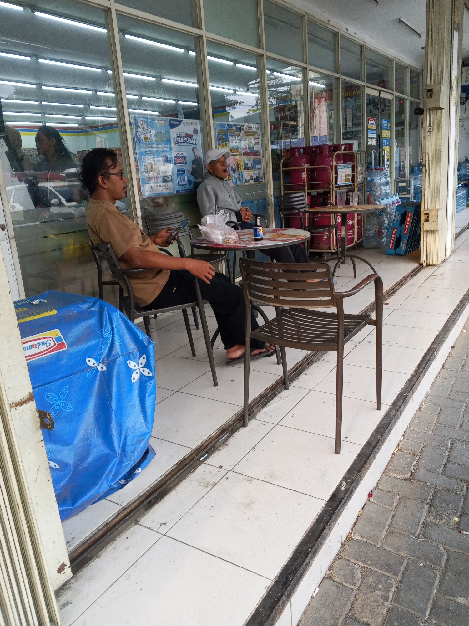 In front of a convenience store in Ciruas, Indonesia, two men sit at small tables on a tiled outdoor area. The man on the left, wearing a tan shirt and black pants, looks at his phone while holding it. The man on the right, in a gray jacket and white cap, appears to be eating or drinking. A blue tarp with a white butterfly pattern covers a large object to the left of the man in the tan shirt. The store's interior, visible through large glass windows, is lined with shelves stocked with various goods, including bottled water and red gas cylinders. The area outside the store is paved with gray bricks, and a metal grate runs along the edge of the tiled area.