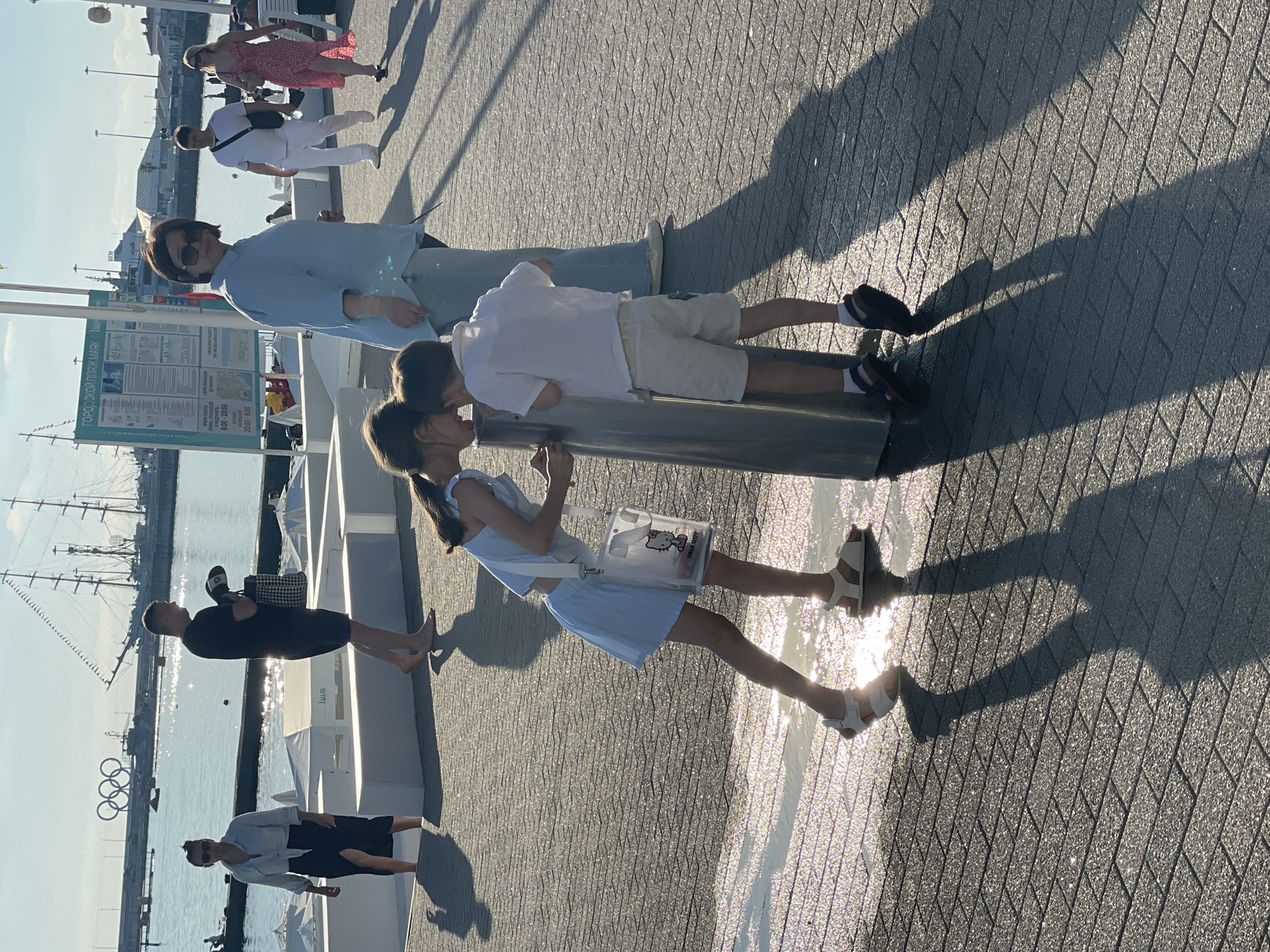 In Sochi, Russia, on a sunny day, two children are drinking from a public fountain on a paved promenade near the sea. The girl, wearing a light blue dress and sandals, leans over the fountain. The boy, in a white shirt and shorts, stands next to her. In the background, several adults walk along the promenade. The sea is visible behind them, with sailboats and ships docked in the harbor. The Olympic Rings are also visible in the distance. The promenade is made of grey paving stones and is wet in some areas, reflecting the sunlight.