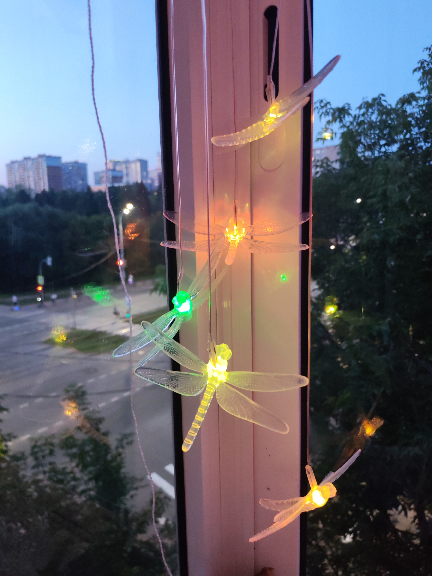The image depicts a window with clear plastic dragonfly-shaped string lights hanging on the inside. The lights are illuminated in various colors, including green and yellow, with some appearing to be orange. Outside the window, it is dusk, with a pale blue sky. In the distance, apartment buildings and trees are visible. A street with a traffic light and a few people can be seen. The scene is likely in Izhevsk, Russia, given the architectural style of the buildings and the time of day.
