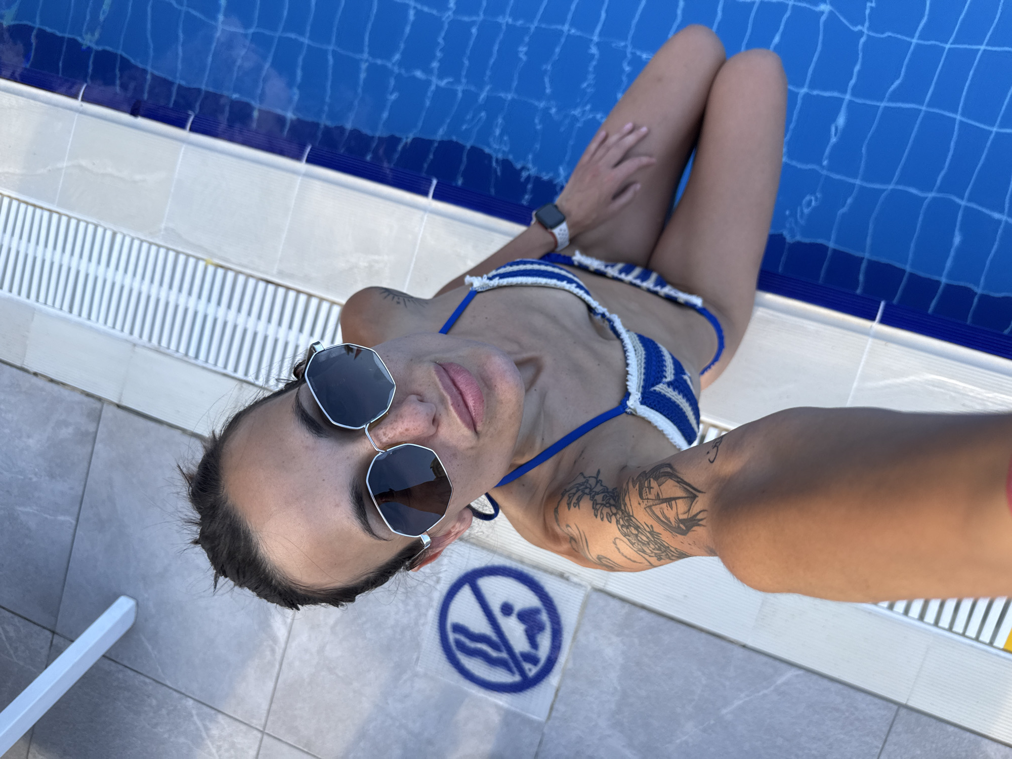 A woman in a blue and white striped bikini and sunglasses sits on the edge of a swimming pool, her legs submerged in the water. Her left arm has a tattoo. A blue and white circular sign depicting a person with a line through them, indicating "no diving," is visible on the tiled ground to her left. The image appears to have been taken in Aksu, Turkey.