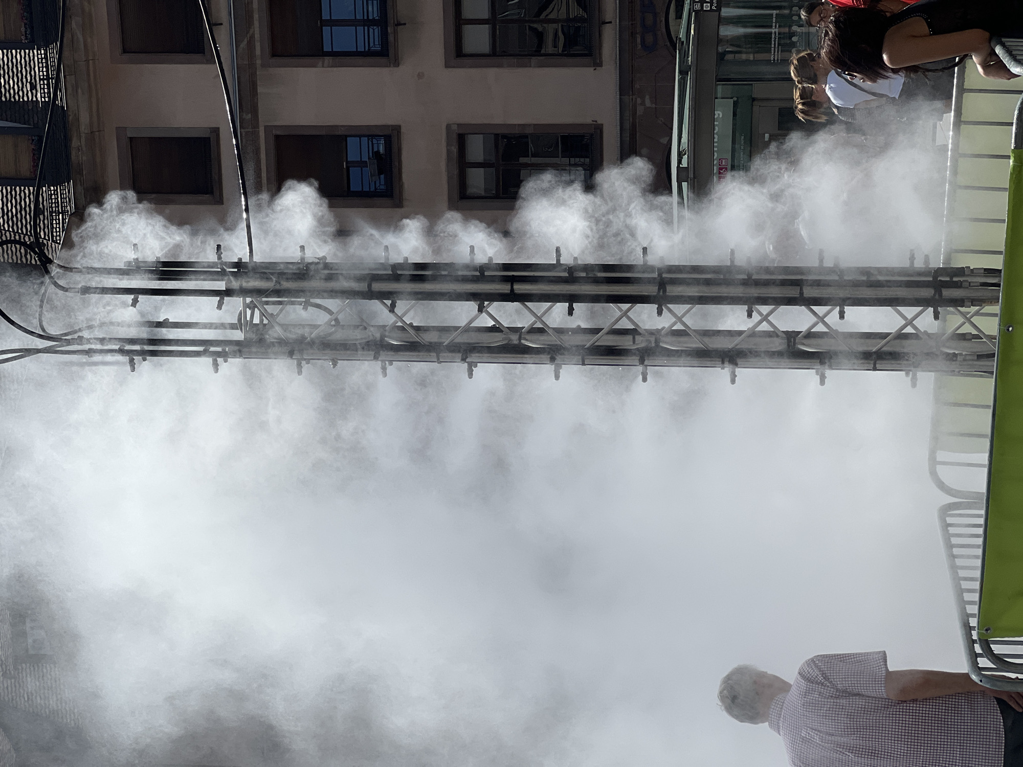 In the city of Unknown, Unknown, a tall metal structure emitting mist stands in the foreground, obscuring much of the scene with a white haze. To the left, a man with graying hair, wearing a checkered shirt, faces away from the camera, looking into the mist. In the background, a building with multiple windows and a tiled roof is visible. To the right, several people are gathered, partially obscured by the mist. One woman in the foreground wears sunglasses and a black top. Behind her, other individuals, including women with ponytails, are visible. A bus stop with signage is also present. Metal fencing with a green barrier runs across the lower portion of the image, suggesting a public space or event. The lighting indicates it is daytime, with bright sunlight contributing to the haziness.