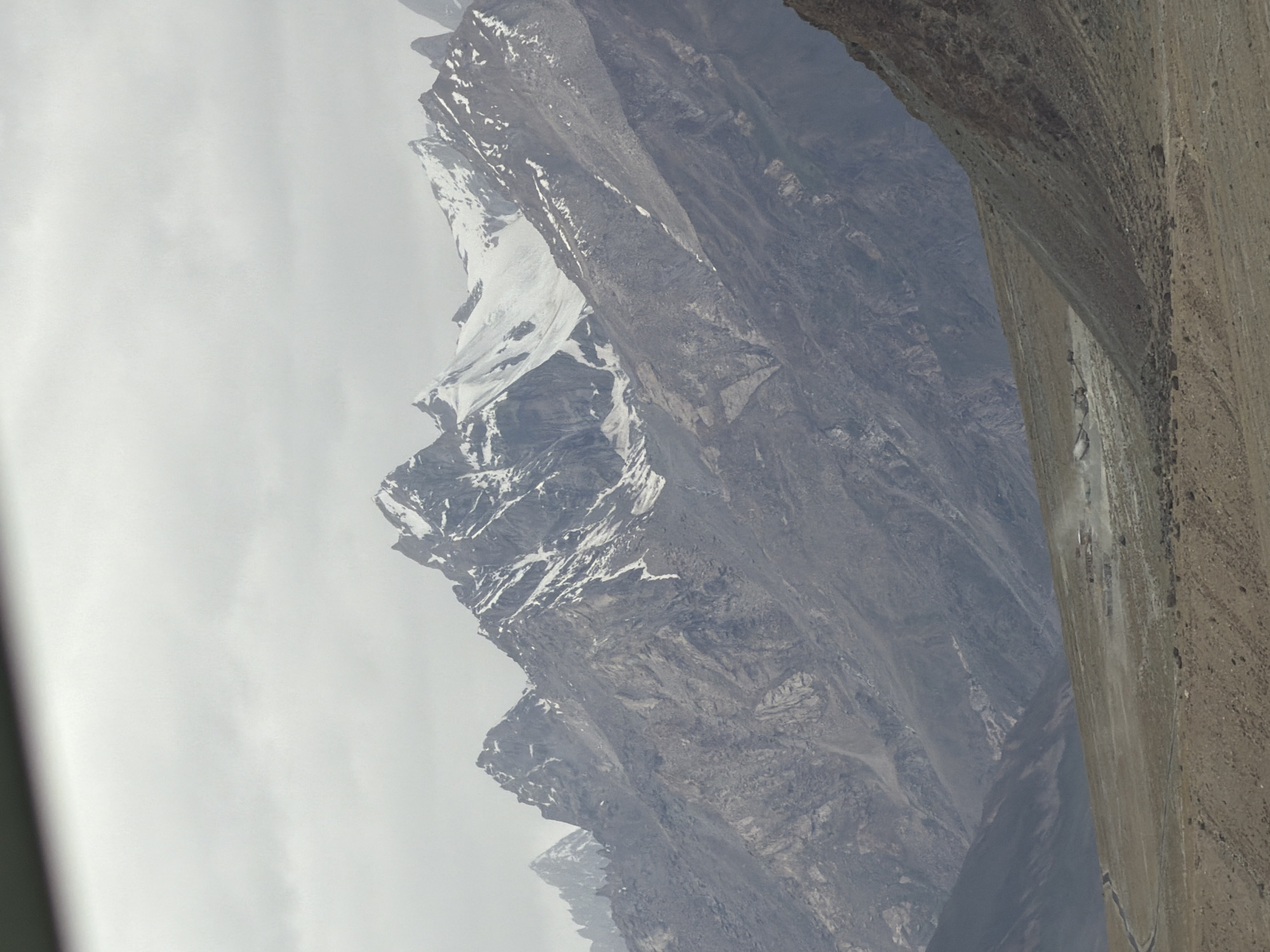 The image depicts a mountainous landscape under a cloudy sky. In the foreground, there is a barren, dusty plain with a winding dirt road. Several small structures, possibly buildings or tents, are visible in the middle ground, with dust plumes suggesting activity or wind. The background is dominated by imposing, rugged mountains with snow-capped peaks. The overall scene conveys a sense of remote, high-altitude terrain. The location is identified as Stongde, India.