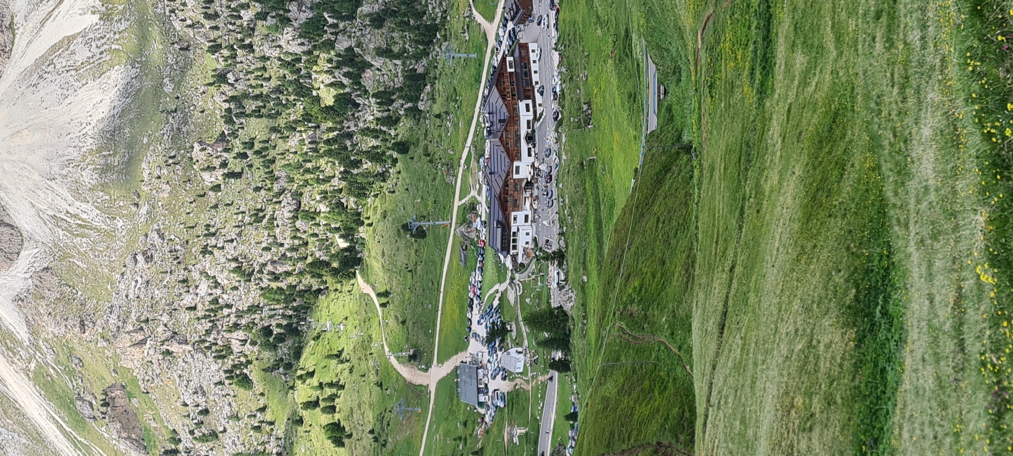 The image shows a panoramic view of a mountain landscape in Selva - Wolkenstein - Selva di Val Gardena, Italy. The scene features a large building complex, likely a hotel or resort, with multiple stories and a dark-tiled roof, situated amidst rolling green hills. Several cars are parked in designated areas around the buildings. In the background, the terrain rises steeply with rocky outcrops and scattered evergreen trees, leading up to a higher mountain peak. Winding dirt paths and ski lift infrastructure are visible on the slopes. The foreground consists of a lush green meadow dotted with small yellow flowers. The sky is not visible in the frame, but the lighting suggests it is daytime.