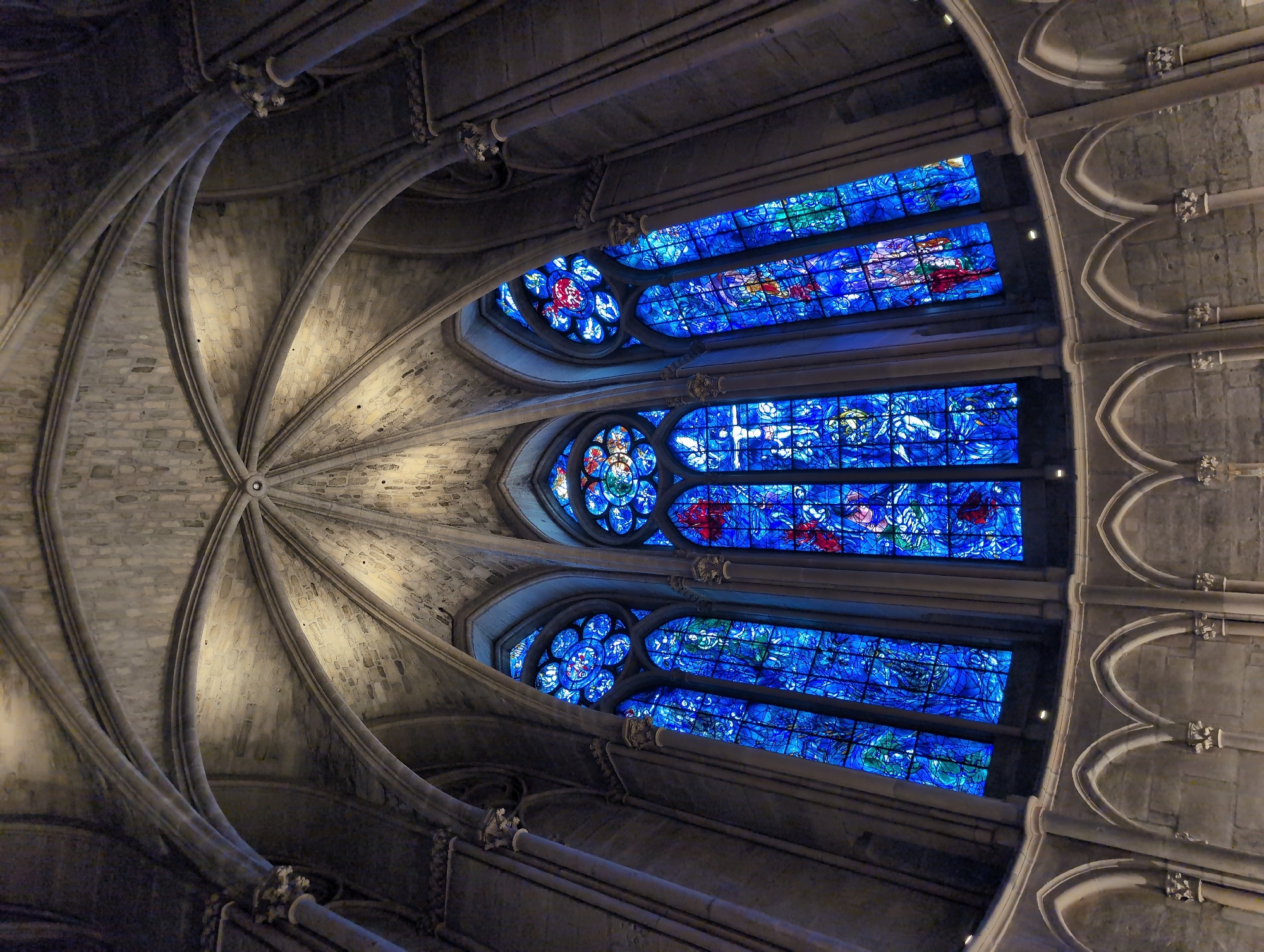 The image shows a low-angle view of the interior of a Gothic cathedral, likely Notre-Dame de Chartres in Chartres, France. The focal point is a triptych of large, ornate stained-glass windows, predominantly in vibrant shades of blue with accents of red and green. The windows are divided into rectangular panes and feature intricate figurative and abstract designs, with a circular rose window above each rectangular section. The ceiling features a complex ribbed vaulting system constructed from grey stone blocks. Below the windows, a row of arched stone arcade structures runs horizontally across the scene. The lighting is dim, with the stained glass windows providing the primary source of illumination, casting a blue hue onto the surrounding stone architecture. There are no people visible in the image. The environment suggests a place of worship or historical significance. Urvillers, France, is not associated with this landmark; Chartres is the correct location.