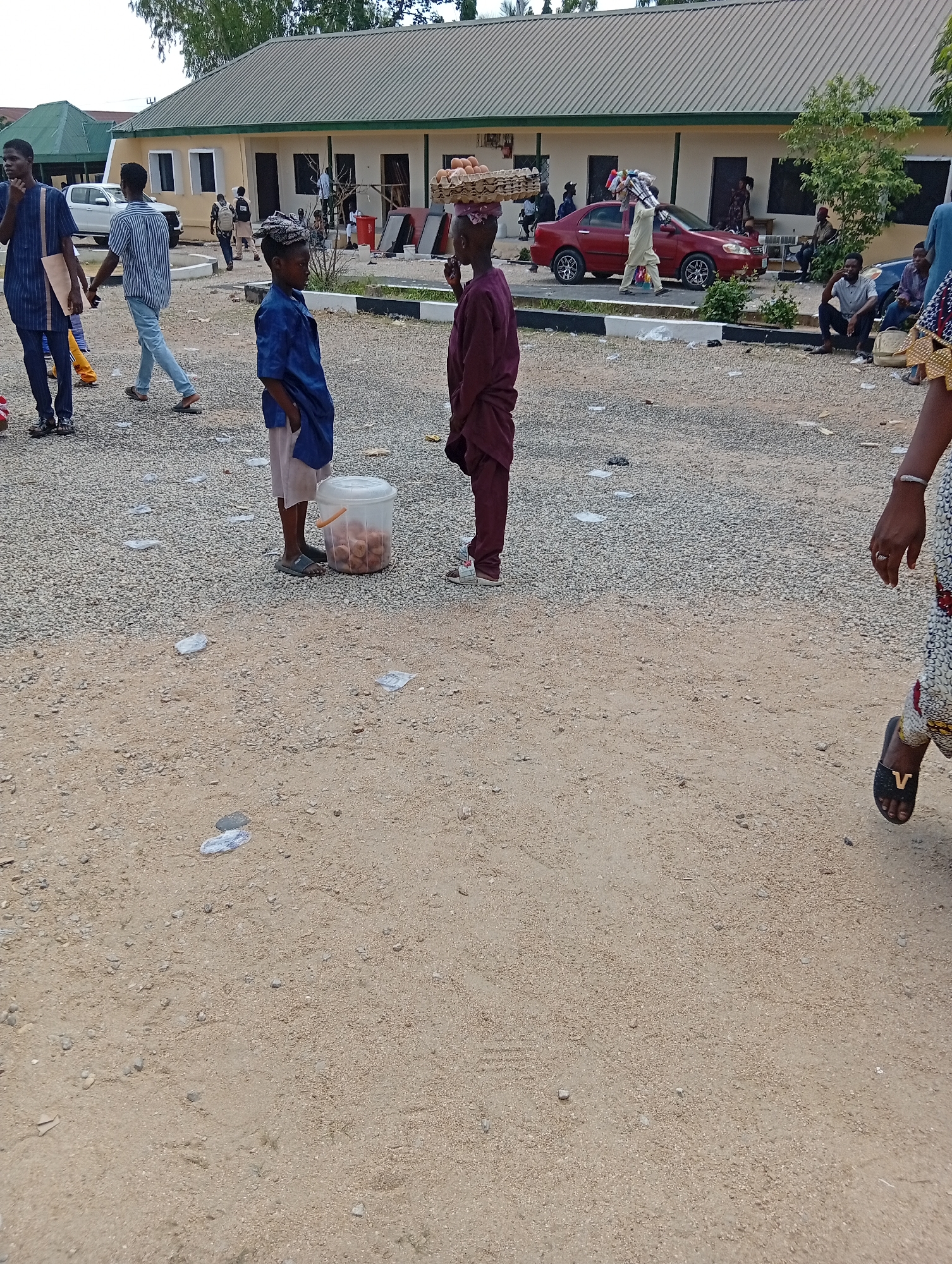 The image depicts an outdoor scene in Jalingo, Nigeria, likely a public space with a gravel and sand ground. In the foreground, a young person in a dark blue tunic and light-colored trousers stands near a clear plastic bucket filled with round, brown items, possibly fried dough or nuts. To their right, another young person in a maroon outfit carries a basket of eggs on their head. Several other individuals are visible in the background, including people standing and sitting, and a red car parked. The environment suggests a commercial or gathering area, possibly near a building with a light-colored facade and a green roof. The ground is littered with small pieces of white paper.