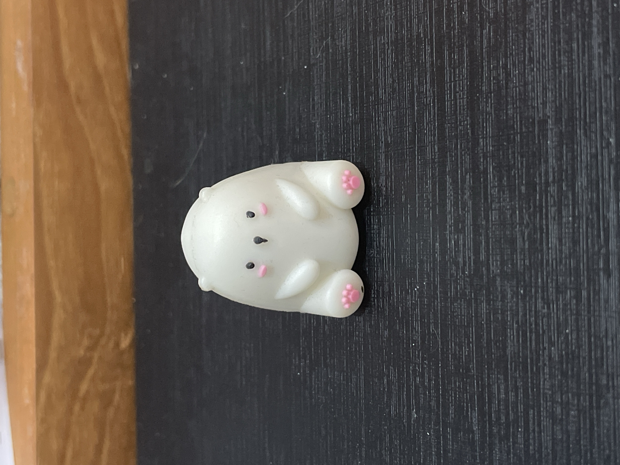 A close-up, eye-level shot shows a small, white, bear-shaped figurine lying on its back on a textured, dark gray surface. The figurine has pink paw prints and facial features. A light brown wooden surface is visible on the left side of the frame. Location information (Denpasar, Indonesia) is not discernible from the visual information provided.