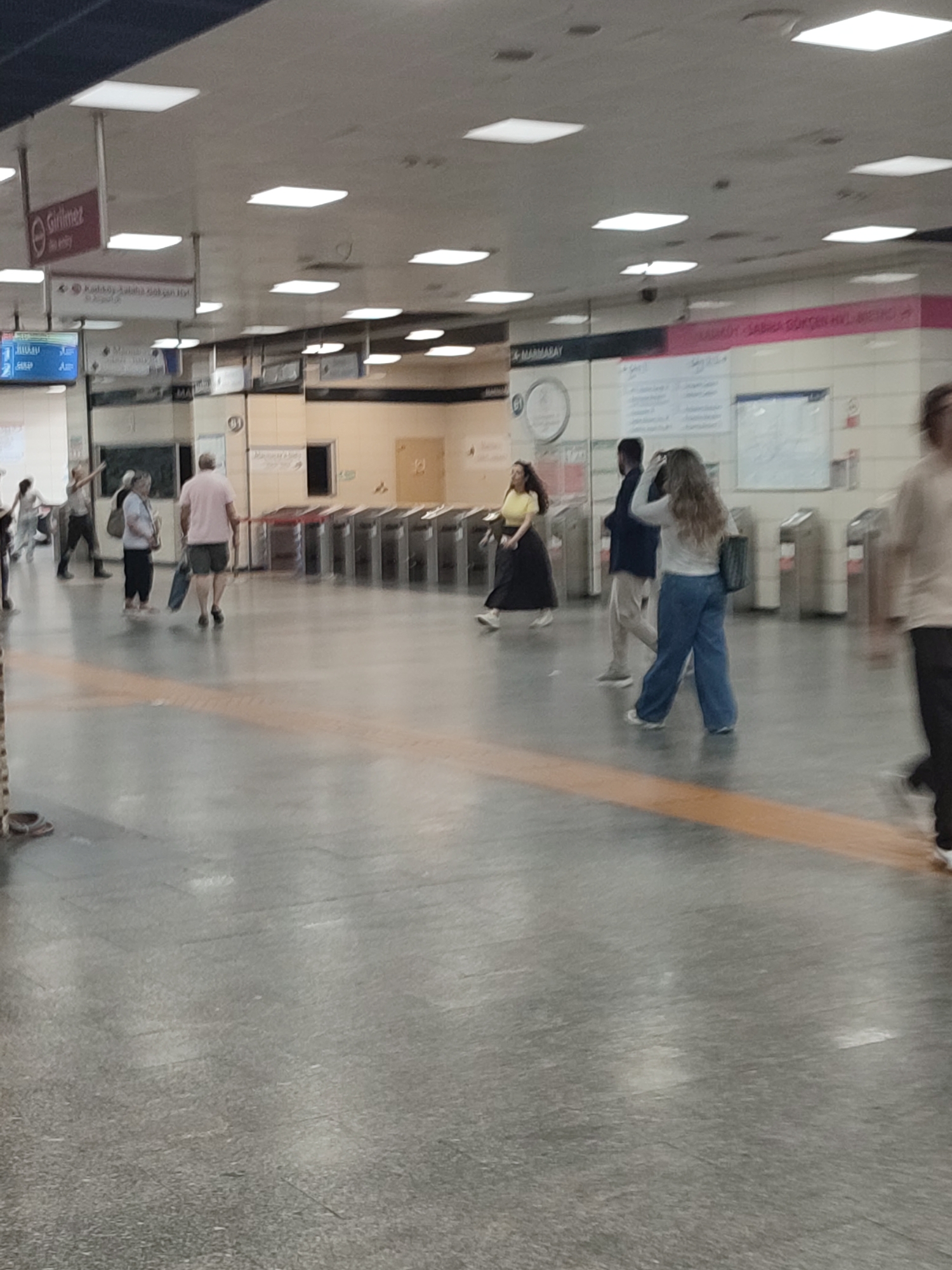 The image captures a scene inside a subway station in Kadıköy, Turkey. The environment is a well-lit, spacious concourse with a polished concrete floor. Overhead, fluorescent lights are visible, contributing to the bright ambiance. Various signs are suspended from the ceiling, indicating directions and station names, including "GİRİLMEZ" (No Entry) and "+ MARMARAY".
In the foreground, a few individuals are walking through the station. On the left, a person wearing a pink t-shirt and shorts is walking away from the camera, carrying a blue bag. To their left, a woman in a yellow top and a long black skirt is walking, looking forward. Further to the right, a person in a white long-sleeved shirt and blue wide-leg pants is walking towards the right, with their back partially to the camera, while another person in dark clothing is walking in the same direction. Several people are blurred in the background, indicating movement.
In the mid-ground, automated ticket gates are lined up, with some people passing through them. The overall impression is of a busy public transportation hub. The architecture suggests a modern subway system, consistent with urban infrastructure in a city like Kadıköy.