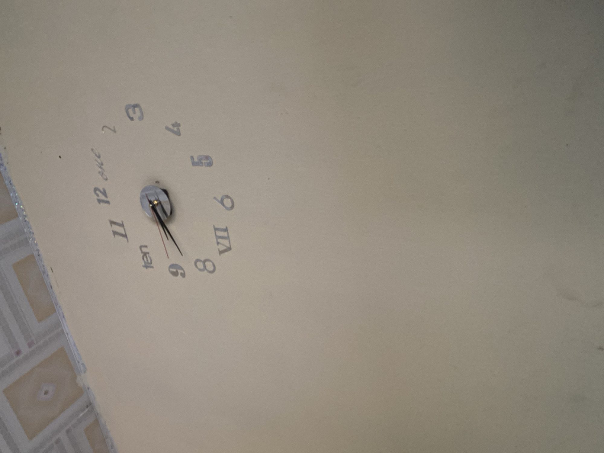 This image shows a close-up of a wall with a clock. The clock has Roman numerals for some hours and Arabic numerals for others, as well as the word "ten" and "one". The hour hand appears to be pointing between 8 and 9, and the minute hand is pointing to approximately the 40-minute mark. The wall is painted a light beige color. Above the clock, the ceiling with a tiled pattern is visible. The scene appears to be indoors. The location is described as Jalingo, Nigeria.