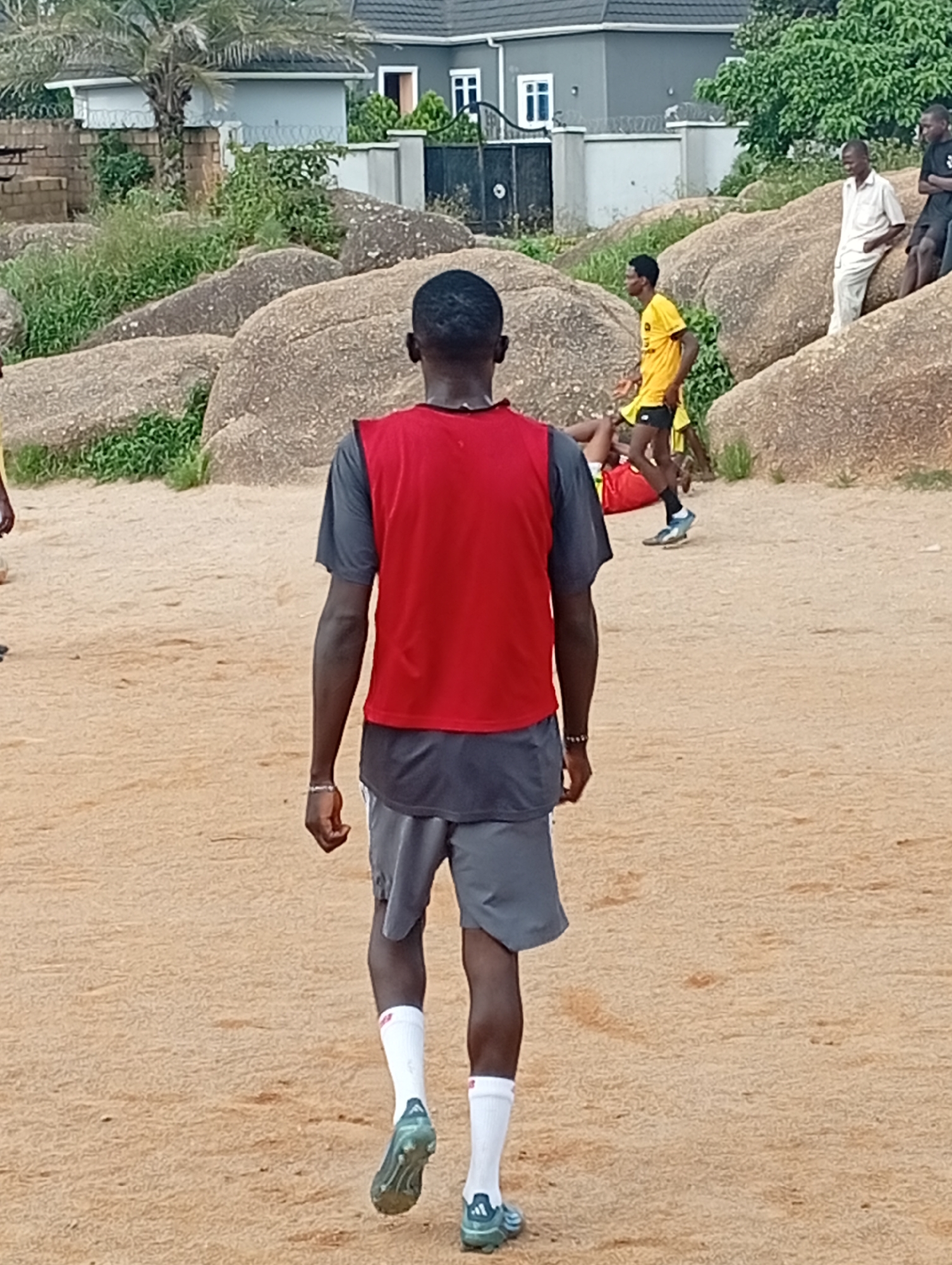 In the city of Jos, Nigeria, the image captures an outdoor scene featuring several individuals engaged in or observing what appears to be a casual soccer activity. The foreground is dominated by the rear view of a person wearing a red vest over a grey t-shirt and grey shorts, with white socks and soccer cleats. This individual is walking away from the camera on a sandy field. In the midground, other individuals are visible. One person in a yellow shirt and black shorts is standing, while another is lying on the ground in a red shirt, possibly as part of the game. Further in the background, two other individuals are seated on large, rounded rocks. One is wearing a light-colored shirt and trousers, and the other is in a black t-shirt and black shorts. Behind the people and the rocky terrain, a modern-looking building with a dark roof and white walls is visible, bordered by green foliage and trees. The overall environment suggests a somewhat rugged, open area, possibly a park or a less-developed part of the city, characterized by sandy ground and large rock formations.
