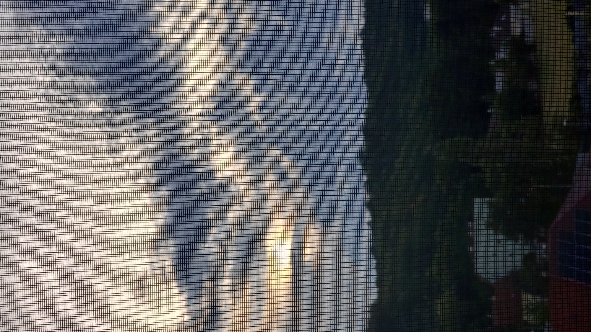 The image shows a view through a window screen of a landscape during what appears to be sunset or sunrise. In the upper portion of the image, a sky with dramatic clouds is visible, with the sun peeking through the clouds, casting a bright, yellowish light. Below the sky is a dark silhouette of a forested hillside, suggesting a natural landscape. In the lower portion of the image, the roofs of several buildings are visible, including one with solar panels. The presence of solar panels suggests a residential or commercial area. Without further information or context, it is impossible to pinpoint the exact location in the city of Prešov, Slovakia. The scene is tranquil and appears to be a typical view from a window in a suburban or semi-rural setting within Prešov.