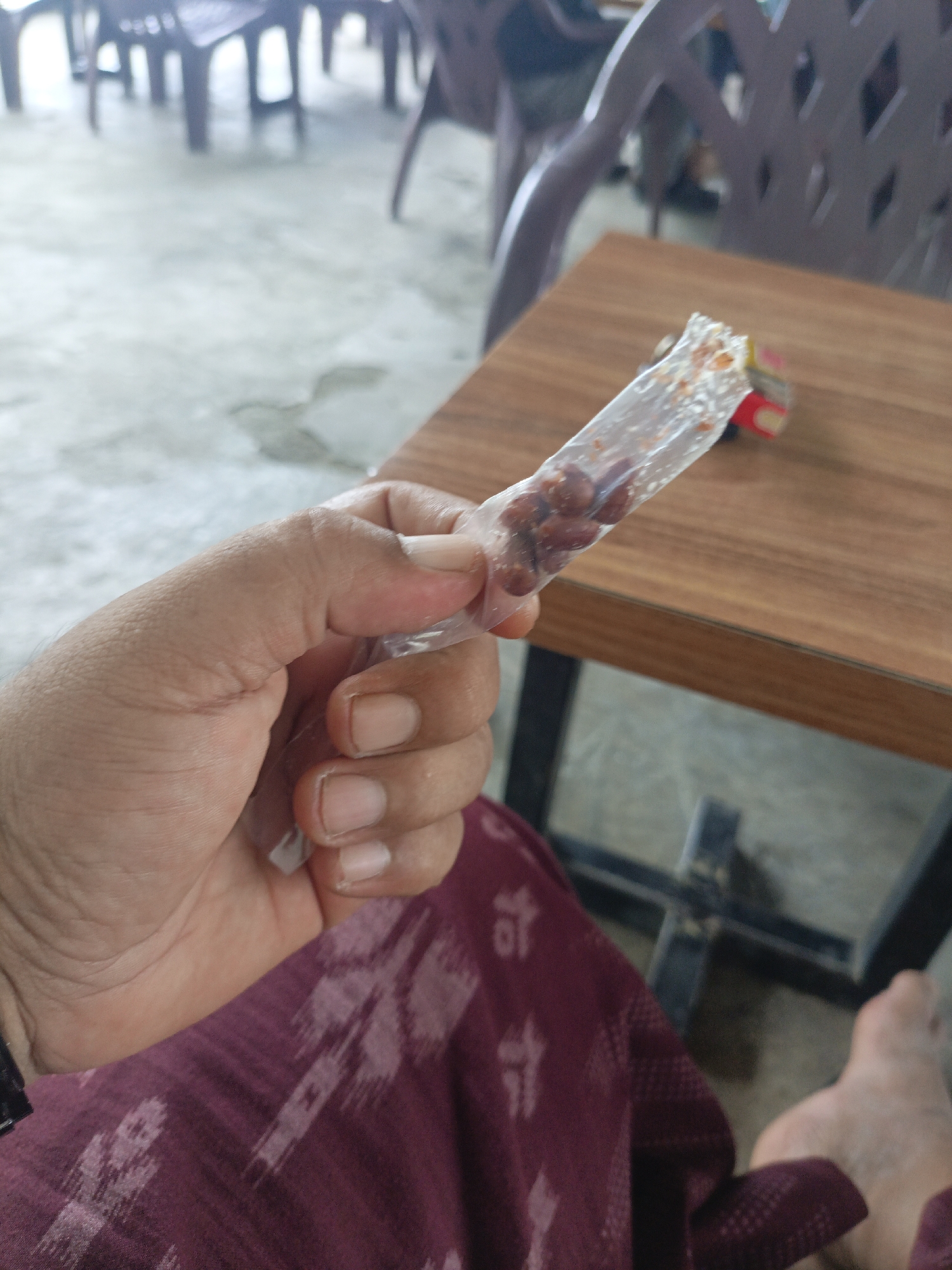 In a casual outdoor seating area, likely a street-side cafe or warung kopi in Lhokseumawe, Indonesia, a person's hand holds a small, clear plastic packet filled with peanuts. The packet is partially unsealed, revealing a portion of the peanuts within. The hand, with visible fingernails, appears to be the right hand of an adult. The person is seated, with a maroon-colored fabric, possibly clothing or a tablecloth, visible in the foreground. Behind the hand and packet, a wooden table with a visible grain is present. In the background, blurry images of dark-colored chairs and a concrete floor suggest the seating arrangement of the establishment. The overall lighting indicates it is daytime.