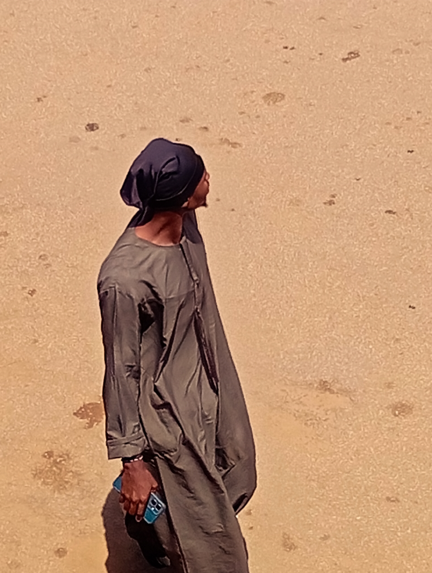 A person is standing in an outdoor area with a sandy or dirt ground. The person is wearing a dark-colored head covering and a long, loose, grey garment. In their right hand, they are holding a blue smartphone. The background is an out-of-focus sandy expanse with some darker specks. The lighting suggests it is daytime.