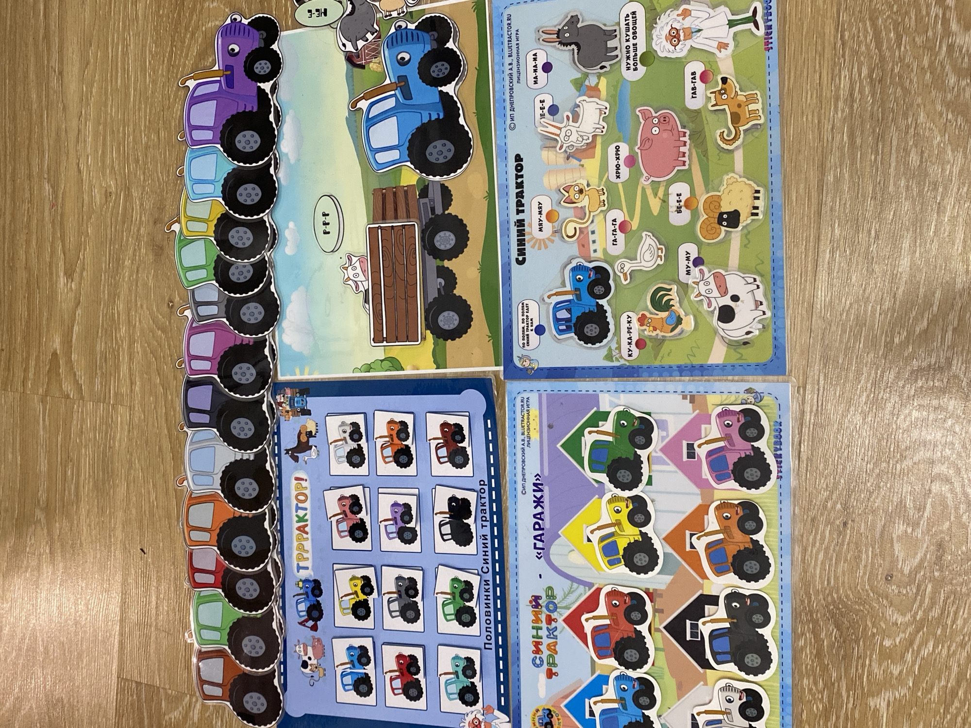 The image shows four learning game boards with cartoon graphics, oriented with their top edges aligned. The boards, which are presumably for children, feature a blue cartoon tractor. The top-left board shows the side of various colored tractors aligned vertically. The top-right board features the blue tractor alongside animal illustrations. The bottom-left board displays tractor halves for matching exercises, and the bottom-right board presents colored tractors to be placed in matching colored garages. The backdrop is a wooden surface. The Cyrillic script on the boards indicates the game is in Russian.