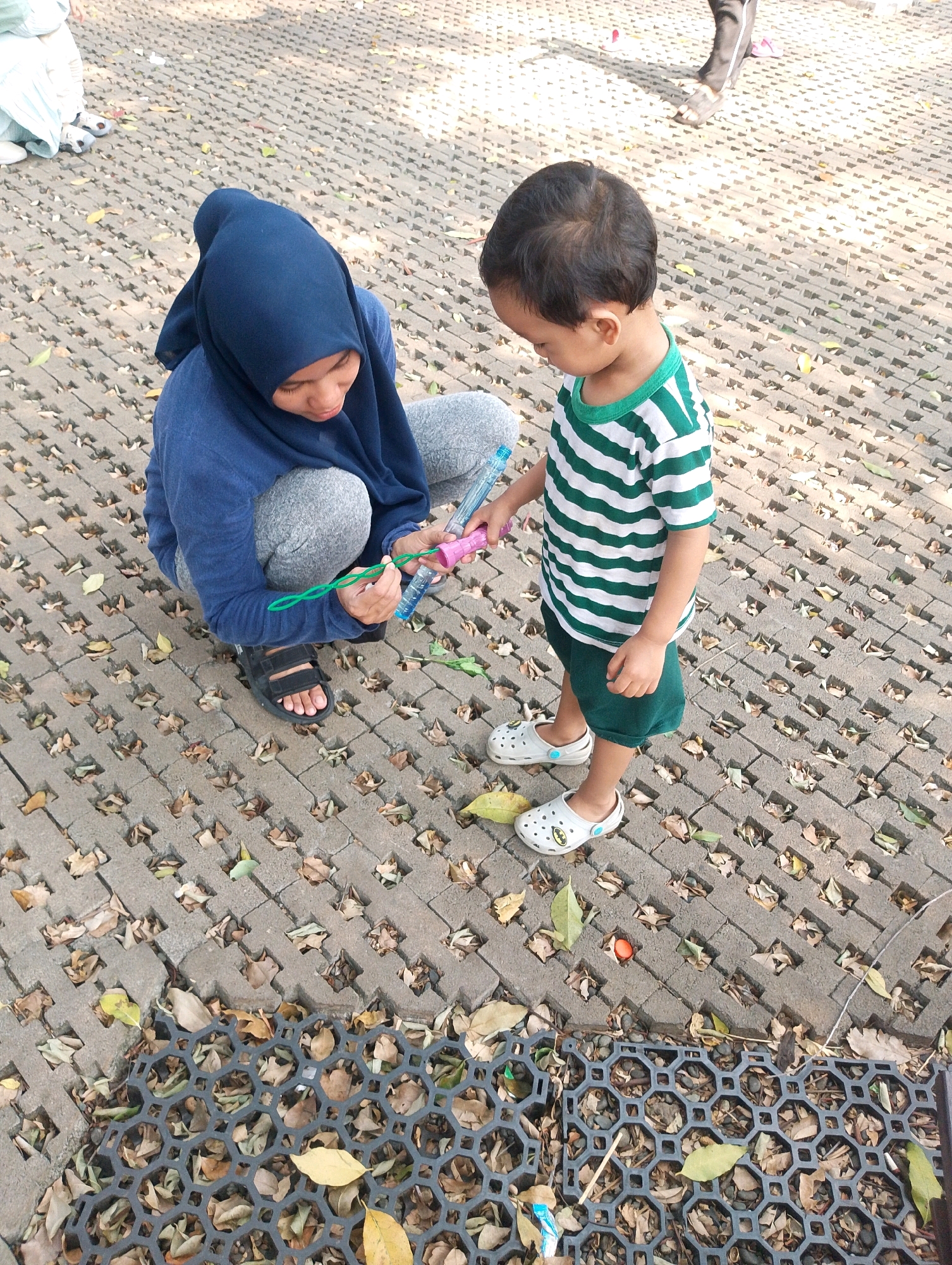 In South Jakarta, Indonesia, on a paved outdoor area with interlocking stone tiles and scattered dry leaves, a woman wearing a dark blue hijab and a blue long-sleeved shirt kneels on the ground. She is holding a green string in her right hand and a pink object in her left, appearing to be a bubble wand and solution. Beside her, a young child, approximately three to four years old, stands wearing a green and white striped t-shirt and dark green shorts, along with white slip-on shoes. The child is looking down at the woman's hands, and the interaction suggests they are engaged in playing with bubbles. The environment is well-lit by sunlight, casting shadows on the ground.