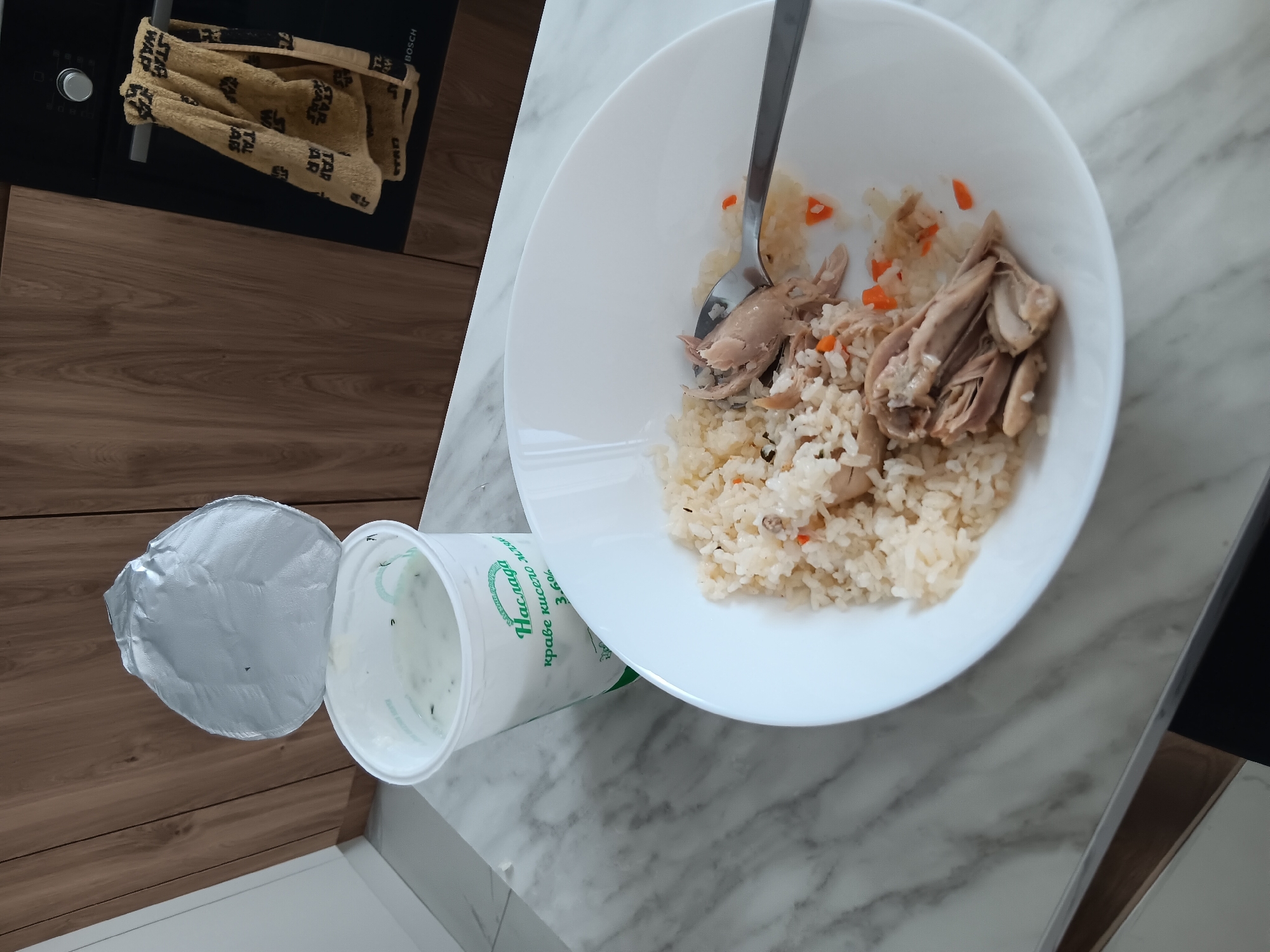 An overhead shot captures a meal set on a modern kitchen countertop. A white plate, partially eaten, holds a serving of rice mixed with what appears to be shredded chicken and small pieces of carrots. A silver spoon rests within the food. Beside the plate, a white plastic cup, likely containing yogurt or sour cream, is visible with its silver foil lid peeled back. The cup has a green label featuring Cyrillic text, which reads "Наслада" (Nasslada, meaning Delight), "краве кисело мляко" (krave kiselo mlyako, meaning cow's sour milk/yogurt), and "3.6%".
The countertop itself exhibits two distinct patterns: a light grey marble-like surface where the food is placed, and a dark wooden surface extending into the background. In the upper left corner, a dark kitchen appliance, possibly an oven or stovetop, is visible with a silver knob and a "BOSCH" logo. A light brown towel, patterned with what looks like "STAR WAR" text, hangs from a bar in front of the appliance. The scene depicts a casual meal in a home kitchen setting, possibly during the daytime given the ambient lighting. No people are visible.