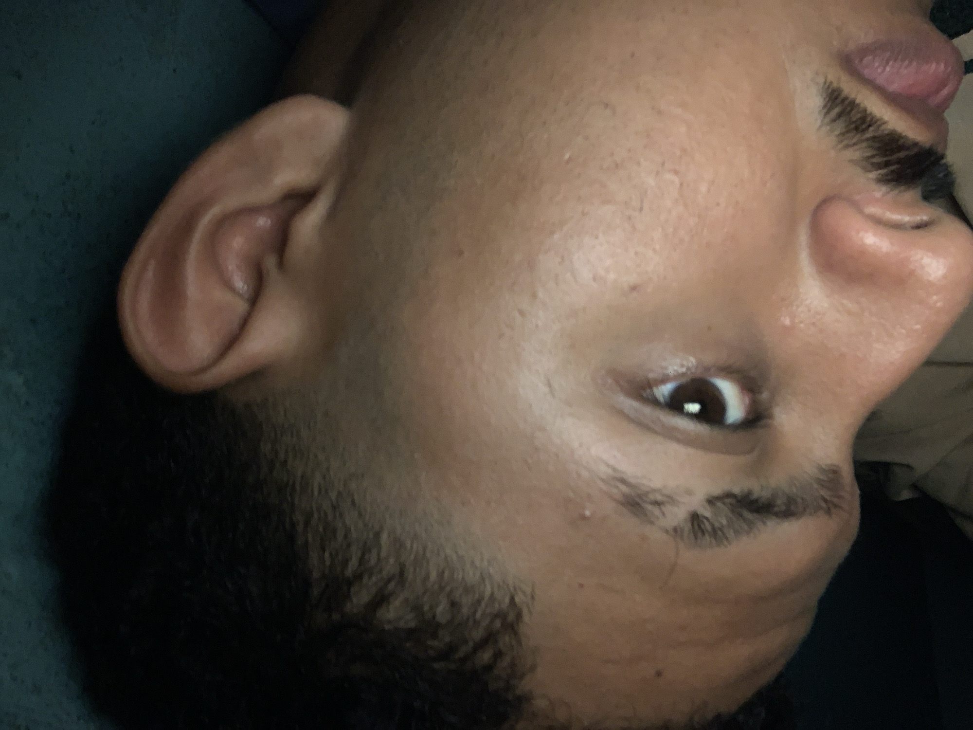 The media shows a close-up, sideways view of an individual's face, likely male, in Santa Ana, Venezuela. The person has a medium to dark skin tone, with visible pores and a slight sheen on the skin, suggesting oiliness or recent hydration.
The visible features include the right ear, part of the cheek, one open eye, a prominent nose, and partially open lips with the tongue visible. The eye is dark brown and appears to be looking directly at the viewer. Dark, short, curly hair is visible on the side of the head, styled with a fade, and there is also some stubble or short facial hair on the cheek and jawline. The eyebrow is thick and dark, with a distinctive shape.
The individual's head is tilted to the left, suggesting they might be lying down or in a relaxed, reclined position. The background is a dark, textured fabric, possibly a blanket or pillow. The expression is largely neutral, possibly indicating a sleepy, relaxed state, or curiosity given the open eye. There is no visible text. The lighting suggests an indoor setting, and the time of day is not discernible. No specific objects are distinctly visible other than parts of the person.