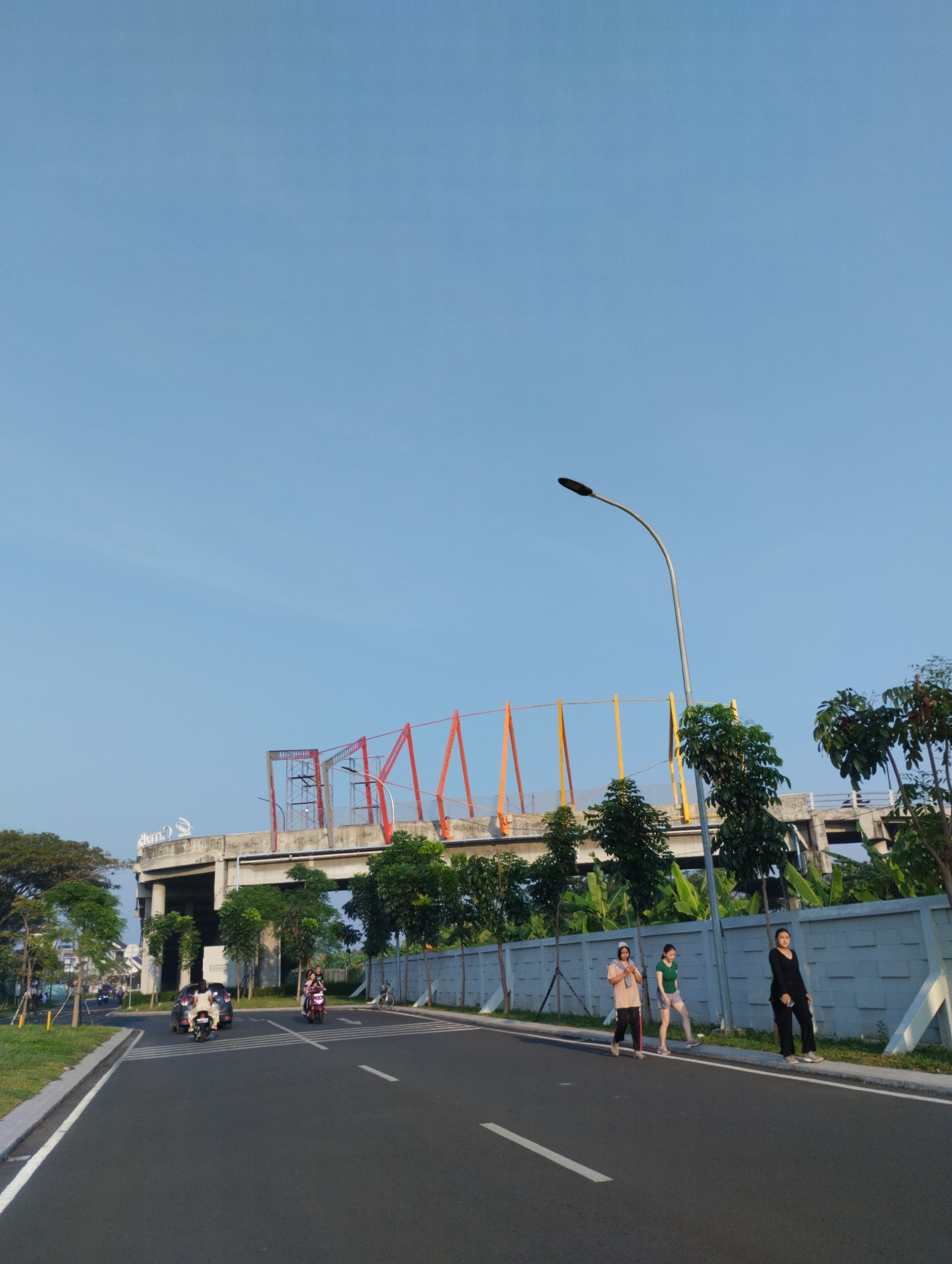 The image depicts an outdoor scene on a paved road in Sidodadi, Indonesia, under a clear blue sky. In the foreground, a black asphalt road stretches into the distance, marked with white lines. On the right side of the road, a low, grey concrete wall with a textured pattern runs along the pavement. Three young women are walking on the grassy verge next to the wall, facing away from the camera. The woman on the far right wears black trousers and a black top. The middle woman wears a green t-shirt and grey shorts. The leftmost woman wears a beige long-sleeved top and dark trousers. Behind them, a line of trees and banana plants are visible.
Further down the road, several motorcycles and a car are traveling. To the left of the road, a grassy area with trees lines the pavement.
In the background, a concrete overpass or elevated structure dominates the midground. This structure features colorful scaffolding and supports, with red, orange, and yellow accents. A large, white, stylized logo, possibly a swan or bird shape, is visible on the top of the structure. A street lamp with a curved pole stands near the road. The overall environment suggests a developing urban or suburban area with infrastructure under construction or recently completed.