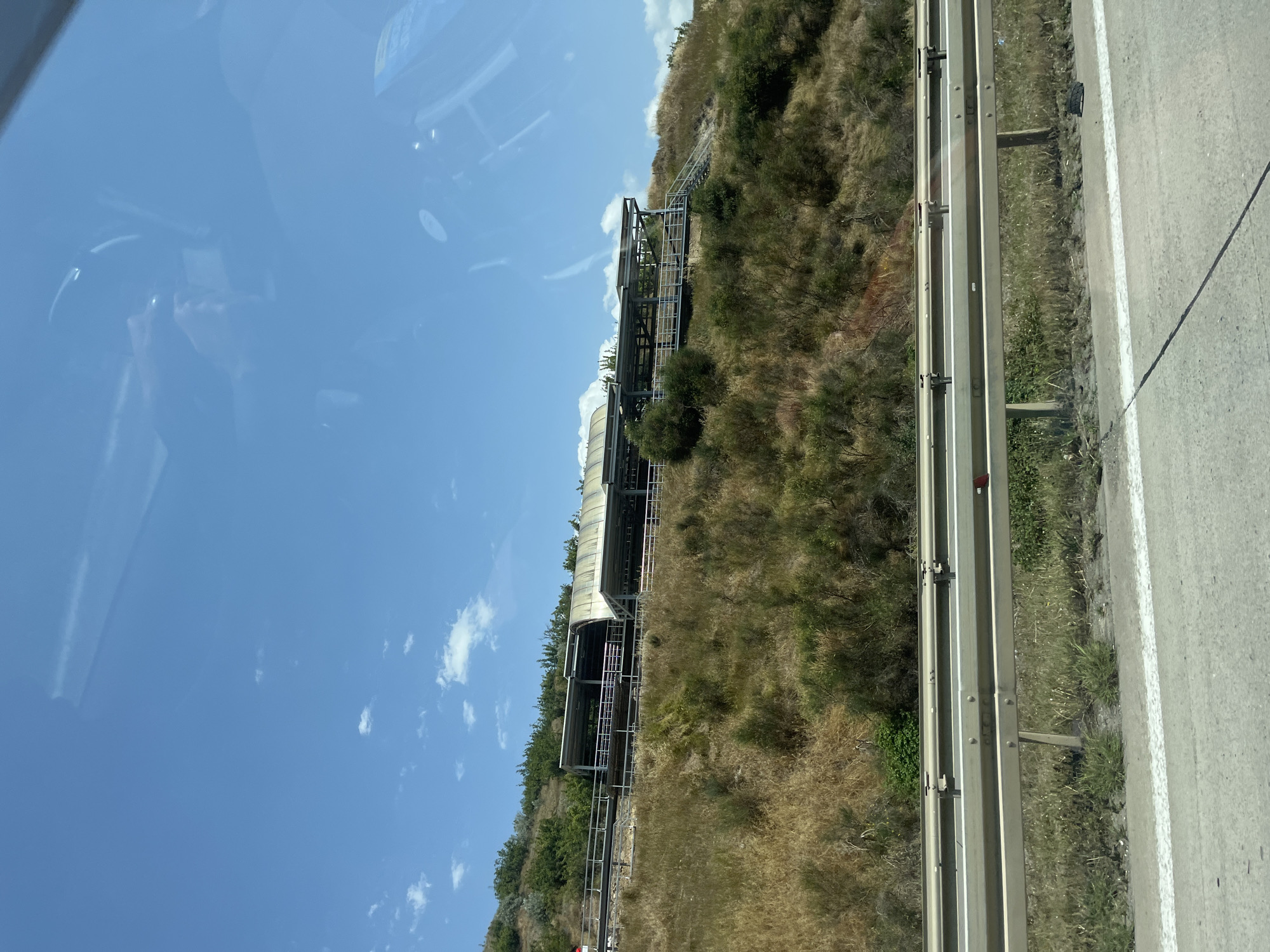 The image depicts a cable car station built into a hillside in Kvemo Rene, Georgia. The station features a long, arched roof structure supported by metal beams and railings. A walkway or platform runs alongside the station, with stairs visible on the right. The hillside is covered in dry, brown vegetation and scattered green bushes and trees. A highway guardrail is in the foreground, separating the road from the hillside. The sky above is blue with scattered white clouds, and there are reflections on what appears to be a car window, suggesting the photo was taken from a moving vehicle.