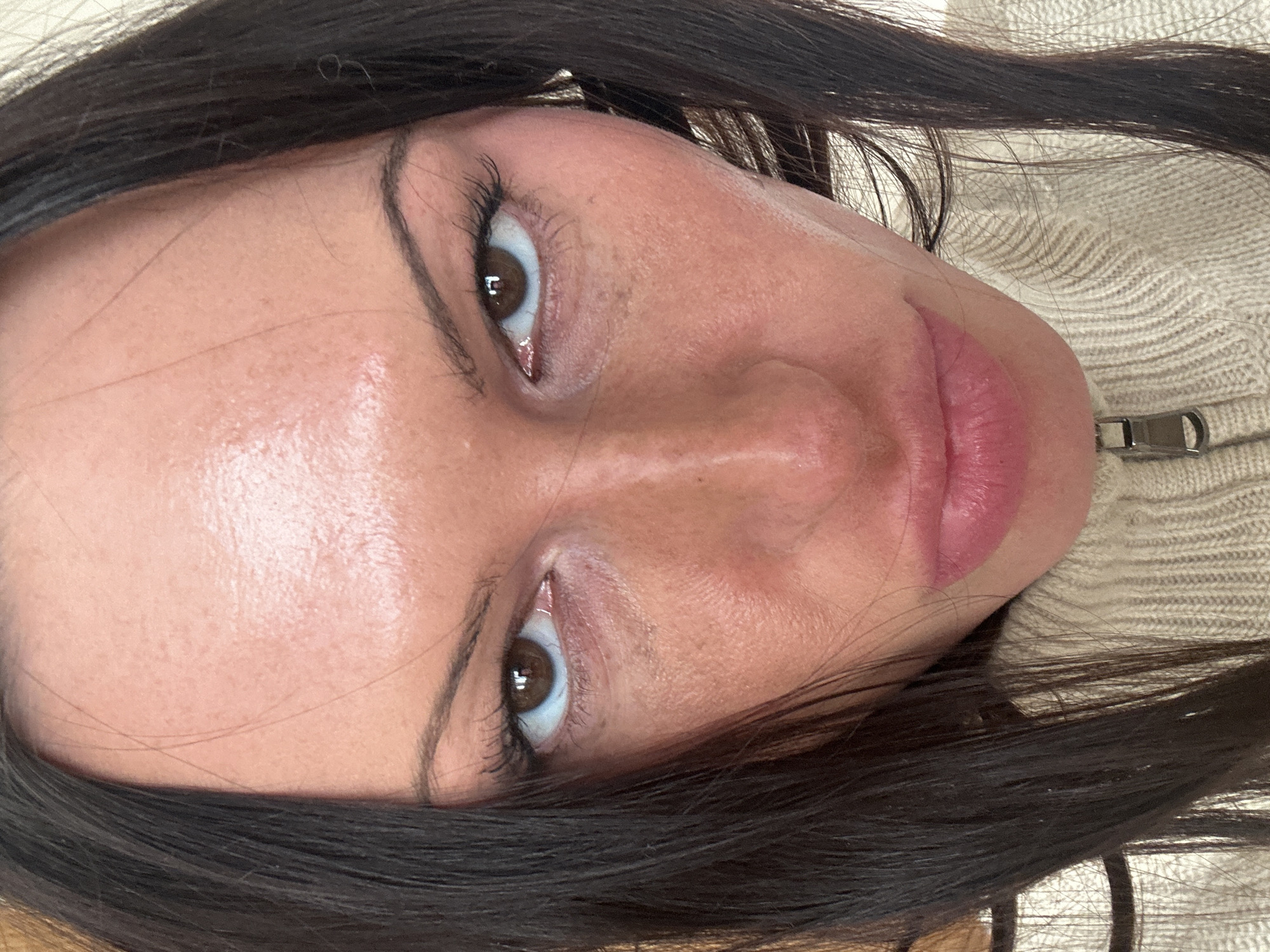 A close-up, angled portrait captures a woman with dark brown hair partially framing her face. Her skin has a dewy appearance. She has brown eyes with visible mascara on her eyelashes. Her lips are a shade of pink. She is wearing a zipped-up, light-colored knit sweater. The location is not identifiable from this image.