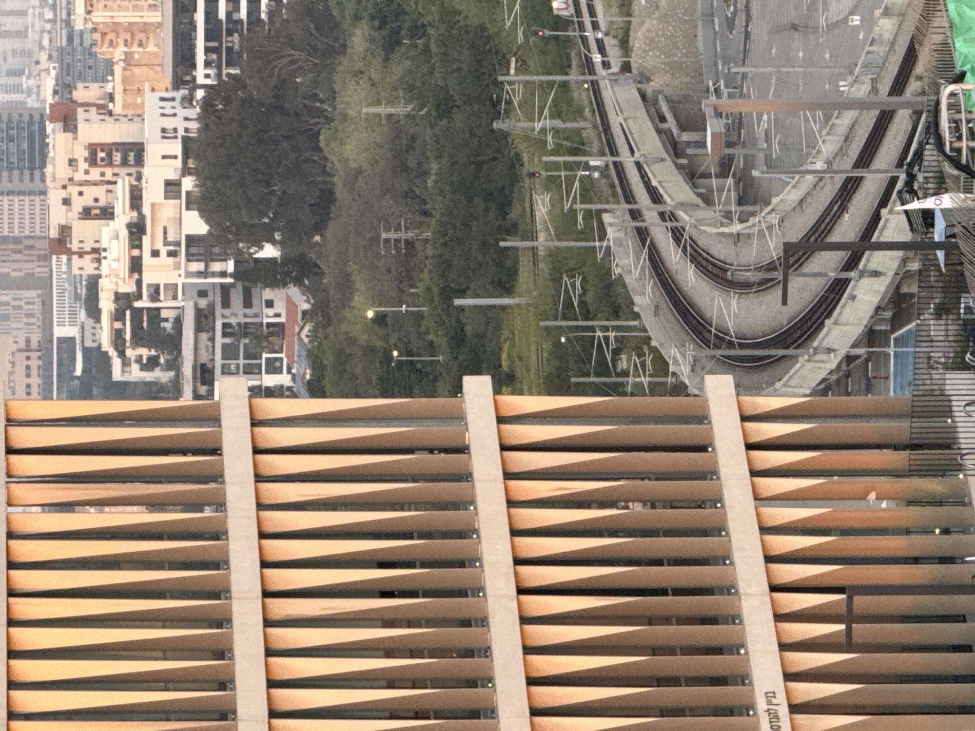 The image shows a view from a building in Tel Aviv, Israel. In the foreground, there is a wooden lattice-like structure, possibly a screen or fence. Beyond the structure is a cityscape with buildings of varying heights and architectural styles. A green area is also visible, with trees and vegetation. Railway tracks curve through the scene. There are electric lines stretching over the tracks, as well as lighting along the tracks.