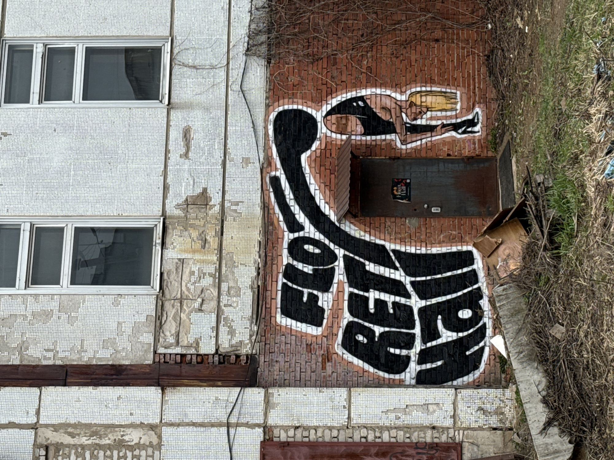 The image depicts the side of a building in Chaikovsky, Russia. The building has a mix of exterior surfaces: a facade covered in small, light-colored mosaic tiles and an adjacent brick wall. Two windows are visible on the tiled section. The brick wall features graffiti: a figure of a woman in a horizontal position with blonde hair, and below that, large black letters spelling out what appears to be a word or phrase in Russian. A dark metal door is partially obscured by the graffiti, and a sticker is visible on the door. Some dead vegetation and debris are present to the right of the brick wall. The building's overall condition suggests neglect and possible disrepair.