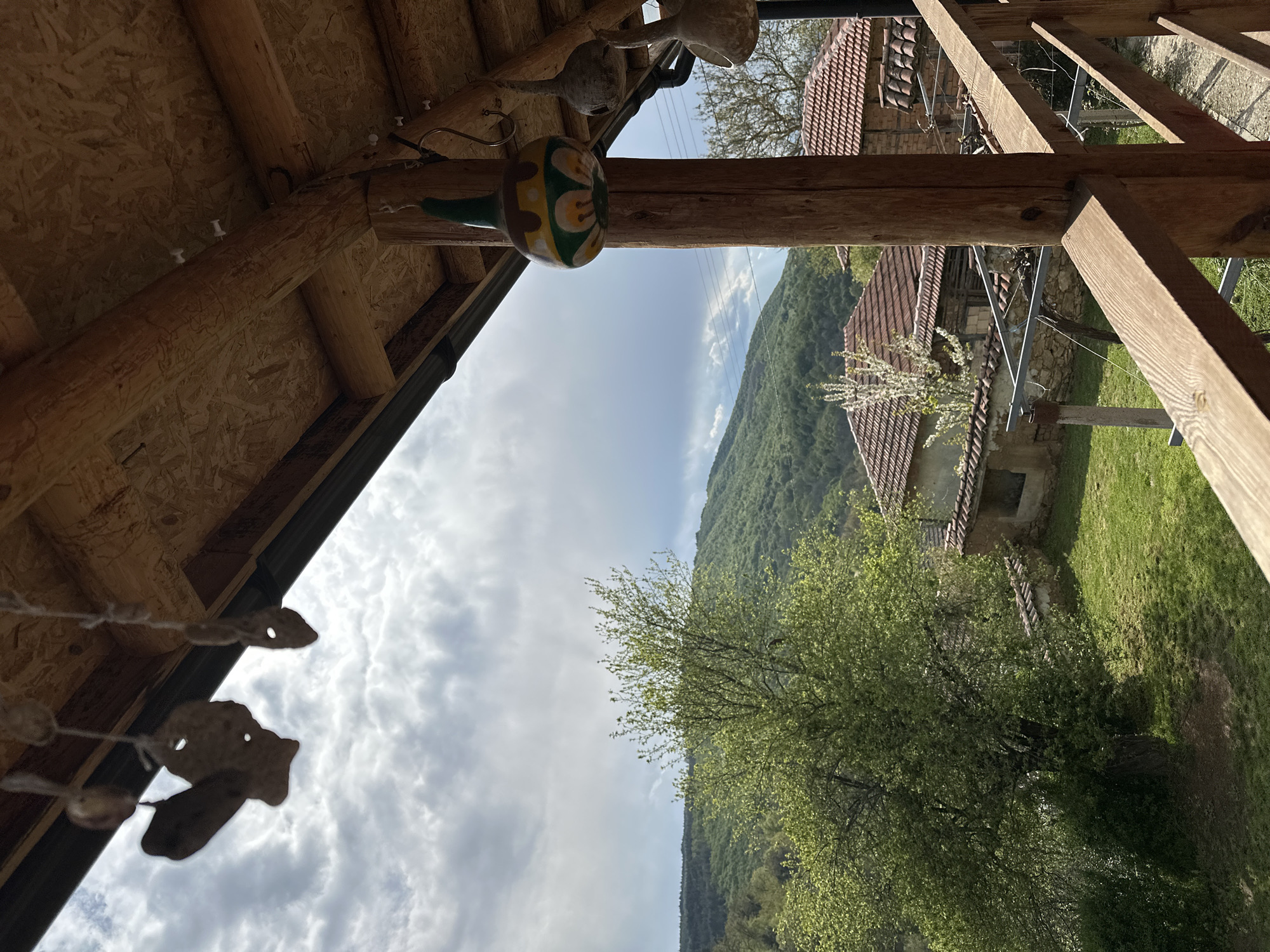 The image, taken from a low angle, shows a view from a wooden structure in what appears to be a rural setting near Mrachenik, Bulgaria. In the foreground, the structure's exposed plywood and wooden beams are visible, with decorative elements hanging from the beams. In the background is a landscape featuring green hills, patches of forest, and a sky filled with clouds. There are also houses with tiled roofs scattered in the middle ground. The lighting suggests it is daytime. No people are visible.