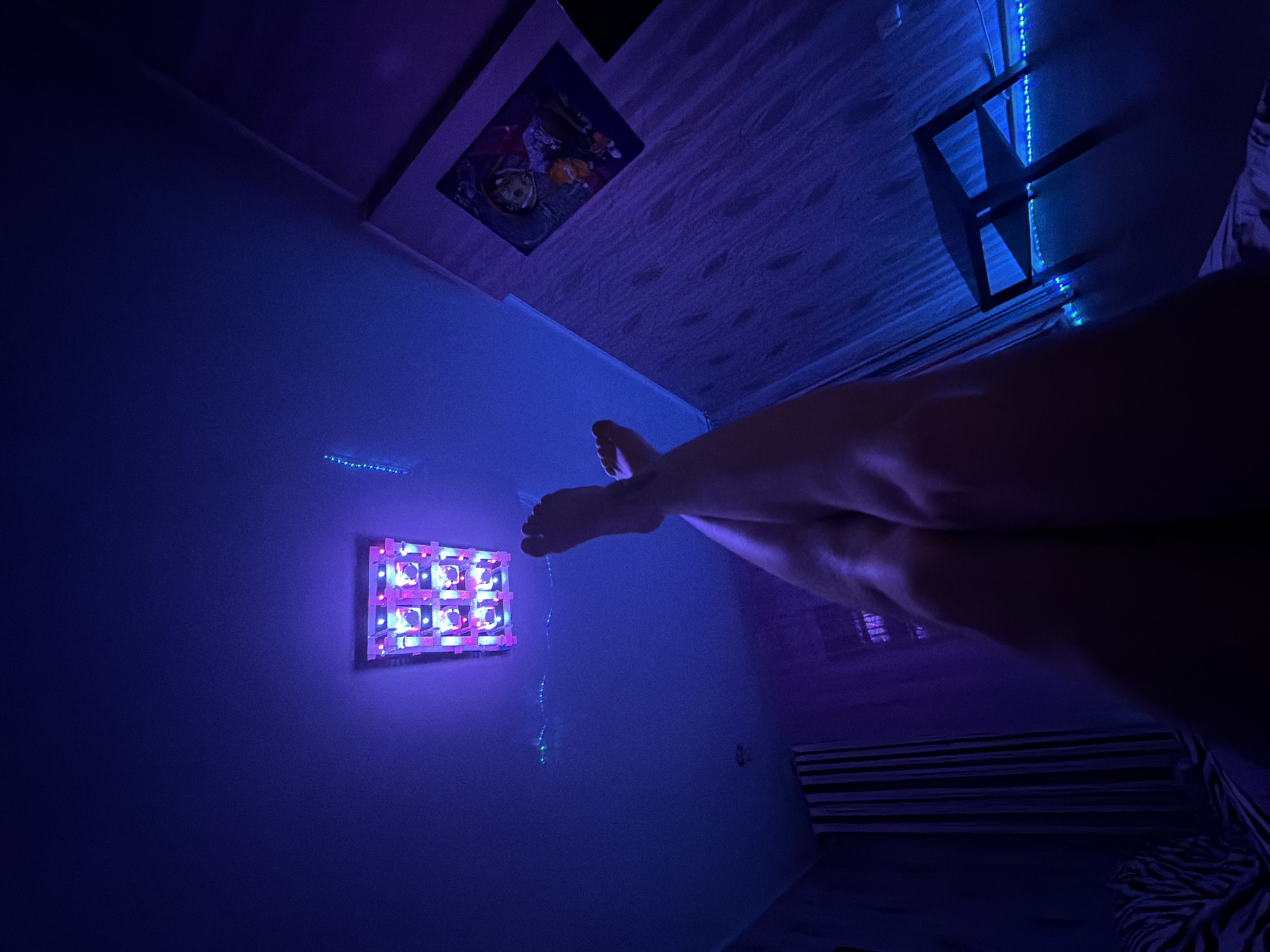 The image depicts a dimly lit room bathed in blue and purple ambient light, likely from LED strip lights and a wall-mounted light fixture. A person's legs and feet are visible in the foreground, positioned as if resting or lounging. The individual's legs are crossed, with the right leg resting on top of the left.
On the wall to the left, there is a rectangular light fixture with multiple small, illuminated squares. This fixture appears to be a type of mood lighting or a specialized grow light. Below it, a string of blue LED lights runs horizontally along the wall. Further up on the wall, a framed picture or poster is visible, featuring cartoonish figures.
In the background, a dark shelf unit with a geometric design is mounted on the wall, also illuminated by blue LED strip lights. The room has striped bedding, suggesting it might be a bedroom or a lounge area. The overall atmosphere is relaxed and intimate, with the lighting creating a cyberpunk or gaming room aesthetic. The time of day is likely evening or night, given the artificial lighting and low ambient light. There is no visible text in the image.