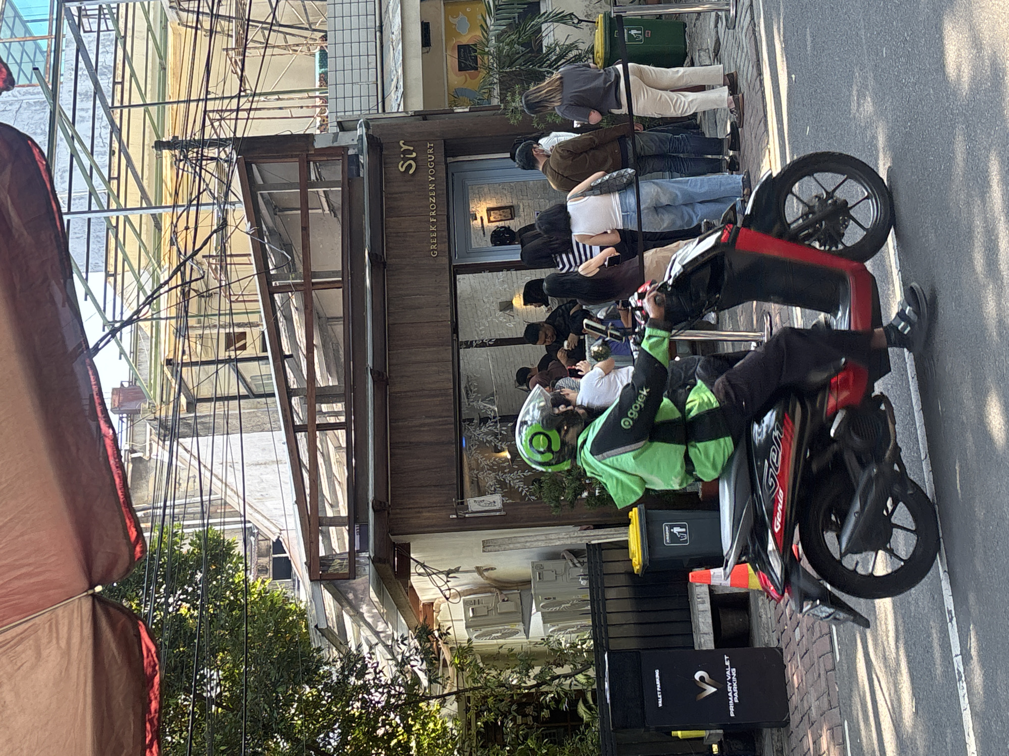 In South Jakarta, Indonesia, a Gojek delivery rider wearing a green jacket and helmet is on a black and red scooter, parked on a street in front of a shop with wooden facade and a sign that reads "Sir GREEK FROZEN YOGURT." A line of people, some wearing masks, are waiting outside the shop. A black valet parking sign is visible on the left, and a yellow-lidded trash bin is next to it. The scene is bathed in sunlight, casting shadows on the road.