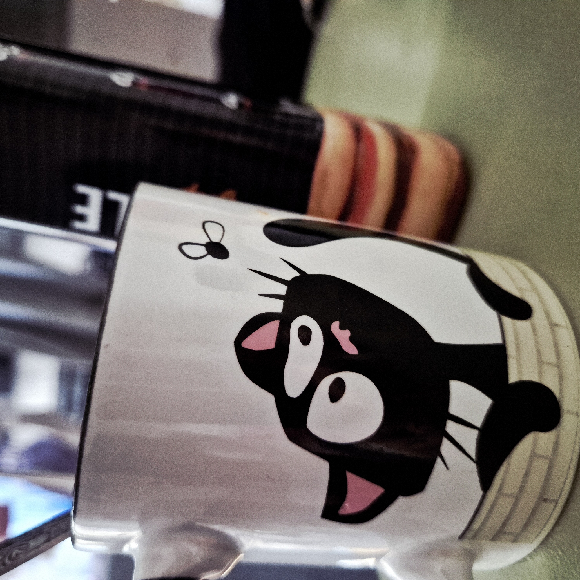 The image shows a white mug with a cartoon of a black cat and a fly printed on it. The mug is lying on its side. The environment surrounding the mug is blurred, with other objects partially visible. Based on my knowledge of Strasbourg, France, I cannot determine the specific location based on this image alone. More visual information of the surroundings would be needed to infer location.