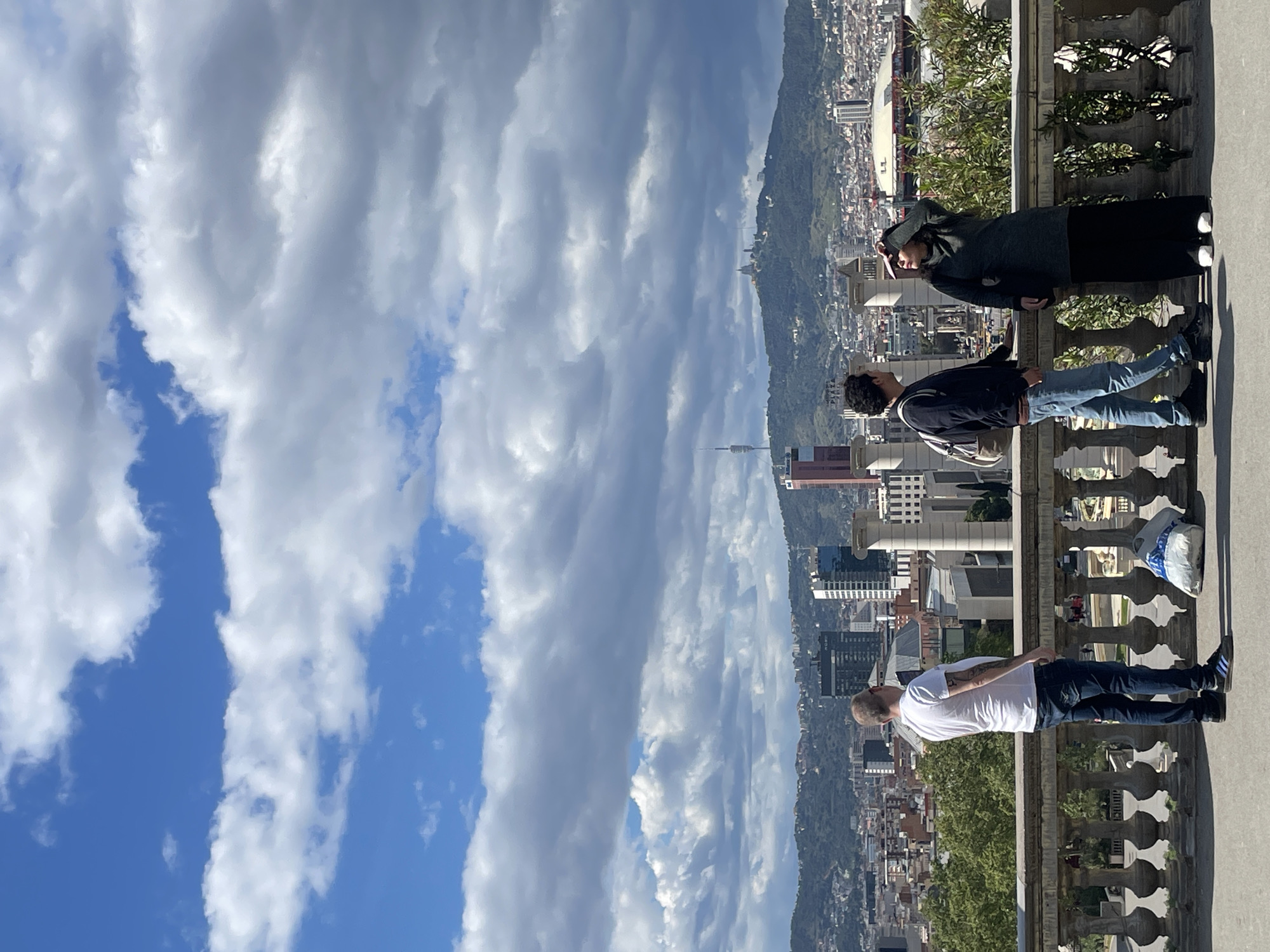 The image shows three people standing on a stone balcony overlooking the city of Barcelona, Spain. To the right of the balcony are buildings and trees, and in the center is a mountain range. A prominent communication tower rises among the city structures. The sky is visible to the left, filled with clouds and patches of blue. The people on the balcony are casually dressed. One is wearing a white t-shirt and jeans, another a black jacket and light-colored jeans, and the third is wearing a dark-colored sweater and skirt. The balcony has a decorative stone railing. A white plastic bag sits next to the man in the white t-shirt. The viewpoint suggests this could be from Park Güell.
