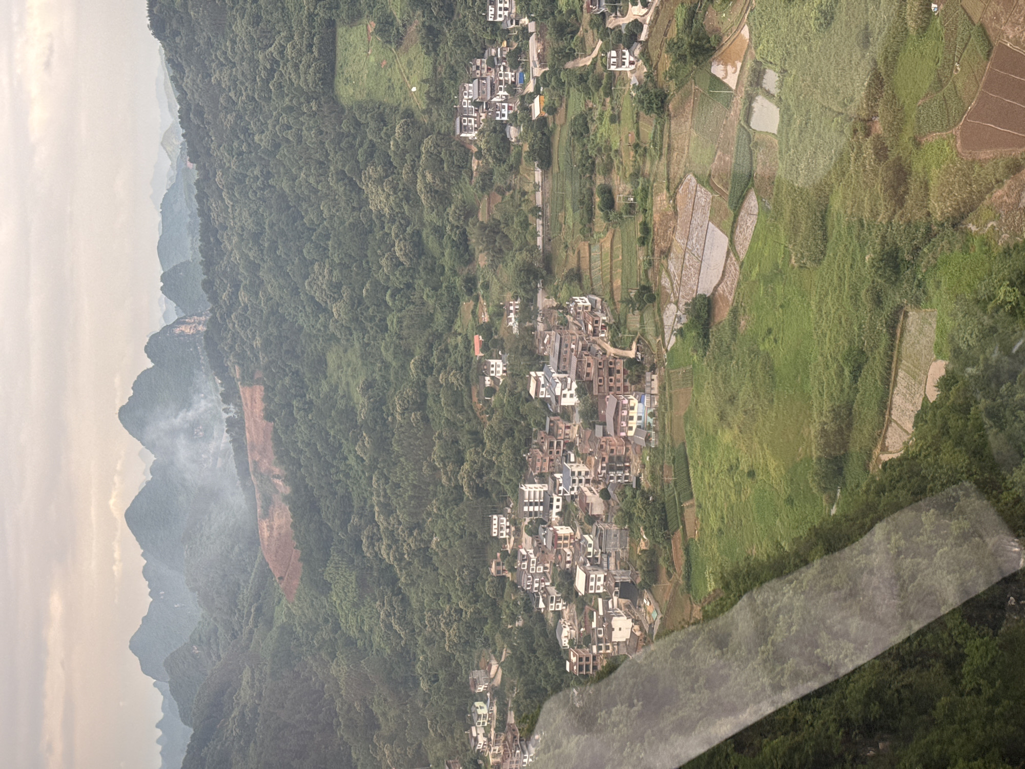 An aerial view reveals a landscape near Guilin, China. The foreground shows lush green fields and terraced rice paddies, partially flooded. A cluster of buildings, possibly a village or small town, is situated amongst trees and fields. In the background, a mountain range appears hazy, suggesting distance and atmospheric perspective. The light is soft, hinting at an overcast day.