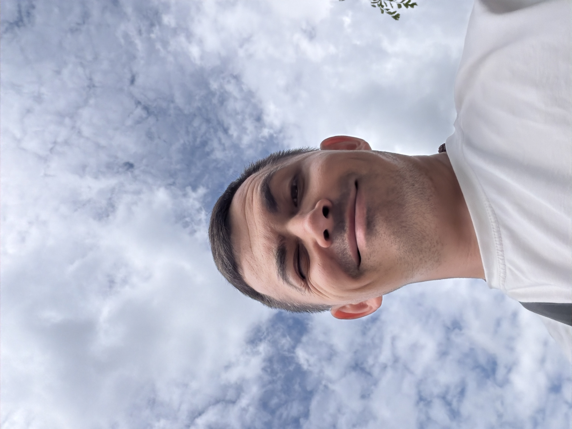 A man with short, dark hair is looking up at the camera, winking and smiling. He is wearing a white t-shirt and a dark strap across his shoulder. The background is a cloudy blue sky. The image appears to have been taken outdoors in Izhevsk, Russia.