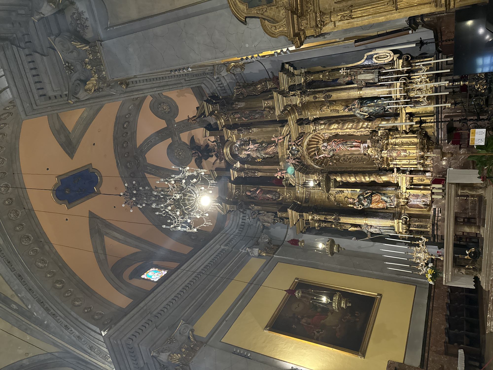 The image shows the interior of a church, likely in Sóller, Spain. The focal point is a large, ornate golden altarpiece featuring multiple statues of religious figures, intricately carved and gilded. Above the altarpiece, a large, crystal chandelier hangs from the ceiling. The ceiling itself is painted with an arched design in a peachy-orange hue, with grey architectural details. To the left of the altarpiece, a large framed painting hangs on a yellow wall, depicting religious figures in muted tones. Several hanging lamps with red tassels are suspended from the ceiling. In the foreground, a white altar cloth covers a table, with a candelabra holding several white candles to the left. Rows of wooden pews are visible in the lower left portion of the image. The overall impression is one of rich decoration and religious solemnity.