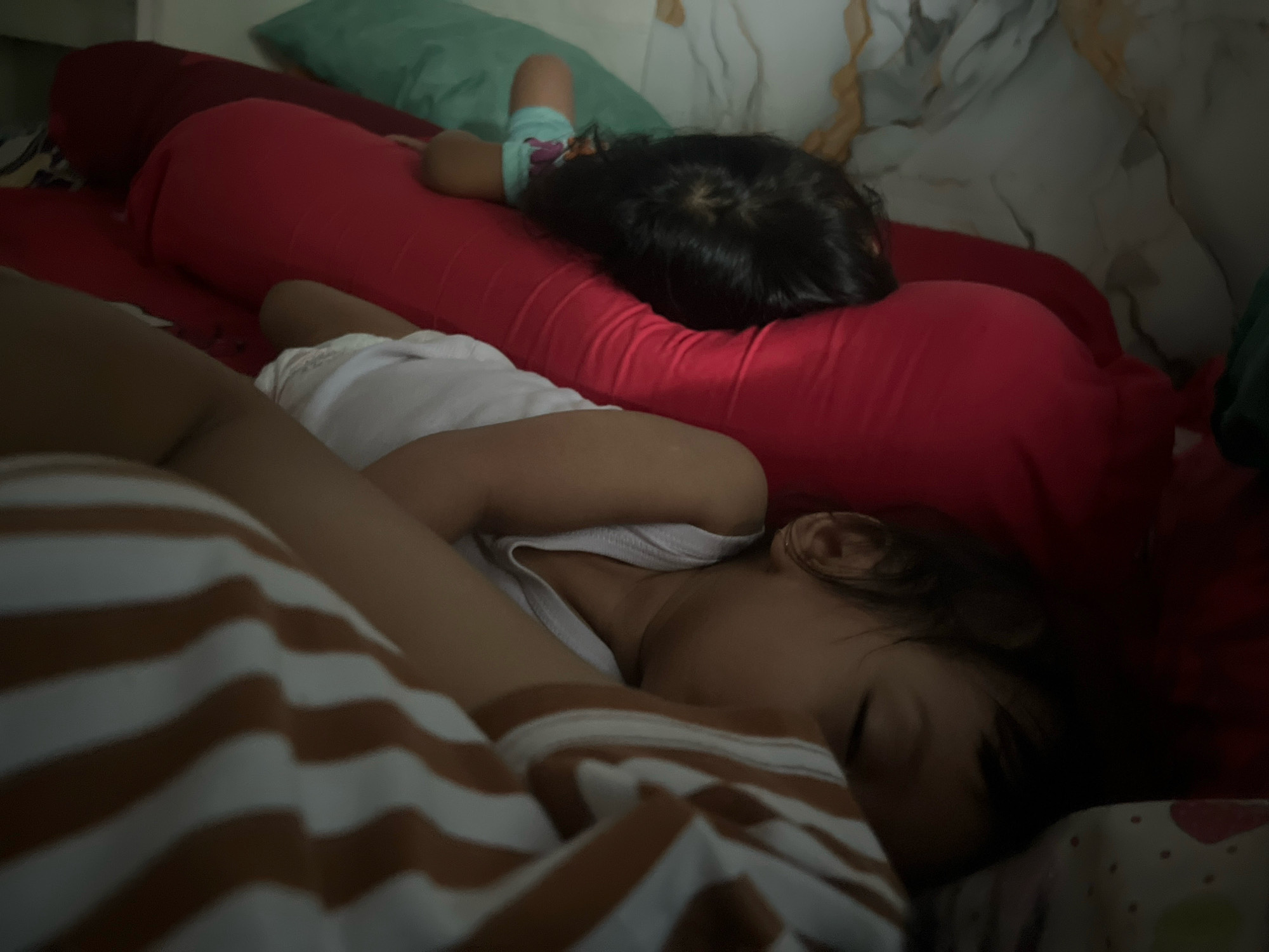 The image depicts a close-up view of two children sleeping in bed, likely in a residential setting in Kecamatan Pati, Indonesia. The child in the foreground is lying on their side, facing away from the camera, with their head on a red bolster pillow and their body draped in a horizontally striped blanket or sheet with brown and off-white stripes. This child is wearing a white tank top. To the left, a portion of another person's arm and shoulder are visible, also in a striped garment. In the background, another child is visible sleeping on their back, propped up on a red bolster pillow, with their head resting on it and their body partially covered by a teal-colored garment. A green pillow is visible behind this child. The overall lighting is dim, suggesting a nighttime or low-light environment. The bedding and pillows appear to be typical household items. The scene conveys a sense of peaceful slumber.
