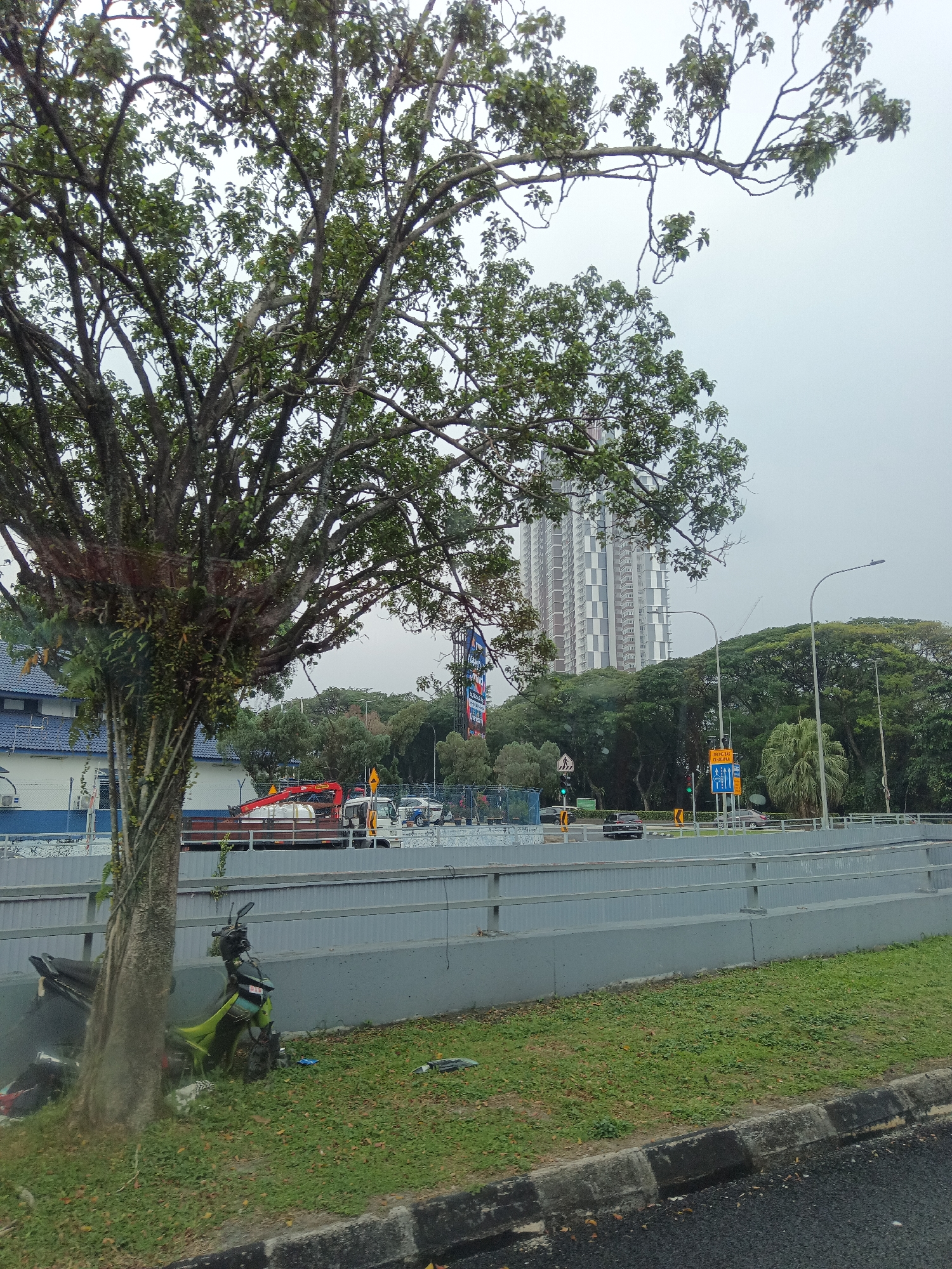 The image captures a section of a multi-level roadway in Kuala Lumpur, Malaysia, on an overcast day. In the foreground, a large tree with dense foliage stands to the left. Below the tree, a damaged green motorcycle is partially visible, resting on the grass. A grey concrete barrier runs along the roadway.
Further back, several vehicles are present on the road. A flatbed truck with a red crane is visible, along with a white truck and other cars. Beyond the road, a tall, modern apartment building with a patterned facade dominates the skyline. A dense canopy of green trees surrounds the building and the roadway. Streetlights and traffic signals are also present. The sky is a uniform light grey, indicating cloudy weather. The scene appears to be a typical urban environment with ongoing traffic and infrastructure.