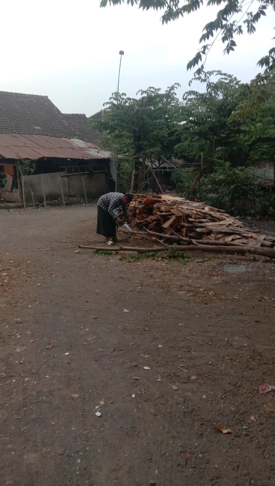 A person is seen from behind, wearing a checkered shirt and a dark sarong, bending over a pile of wood. The individual appears to be using a machete on a fallen branch. The scene is set in an outdoor environment with a dirt ground. In the background, there are structures that appear to be houses with corrugated metal roofs and trees. A tall pole with a dark object at the top is visible in the distance. The overall impression is that of a rural or semi-rural setting, possibly a yard or a roadside in Karang Anyar, Indonesia.