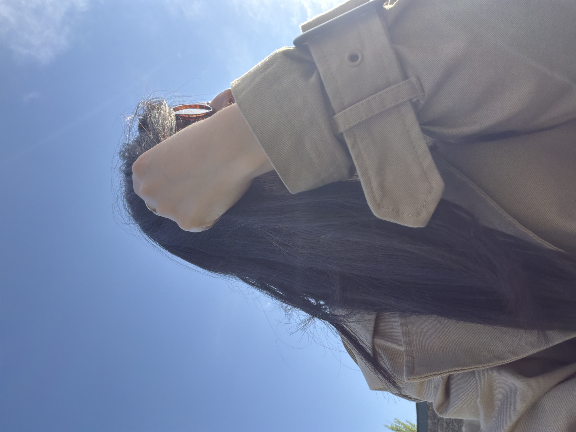 The image captures a partial view of a person looking upwards, set against a bright, clear blue sky. The individual has long, dark hair and is wearing a light beige or khaki trench coat or jacket with visible strap details on the sleeve. A light-skinned hand is visible near their head, with a pair of brown-rimmed sunglasses perched on top of their hair. The predominant feature of the background is a vibrant blue sky, dotted with a few wispy white clouds and a bright sun, causing a noticeable lens flare near the top. The light suggests it is daytime with direct sunlight. A small area of green foliage and a stone-like structure are barely visible at the bottom right, indicating an outdoor setting, potentially in Yerevan, Armenia. The scene conveys a sense of open air and pleasant, sunny weather. No visible text is present.