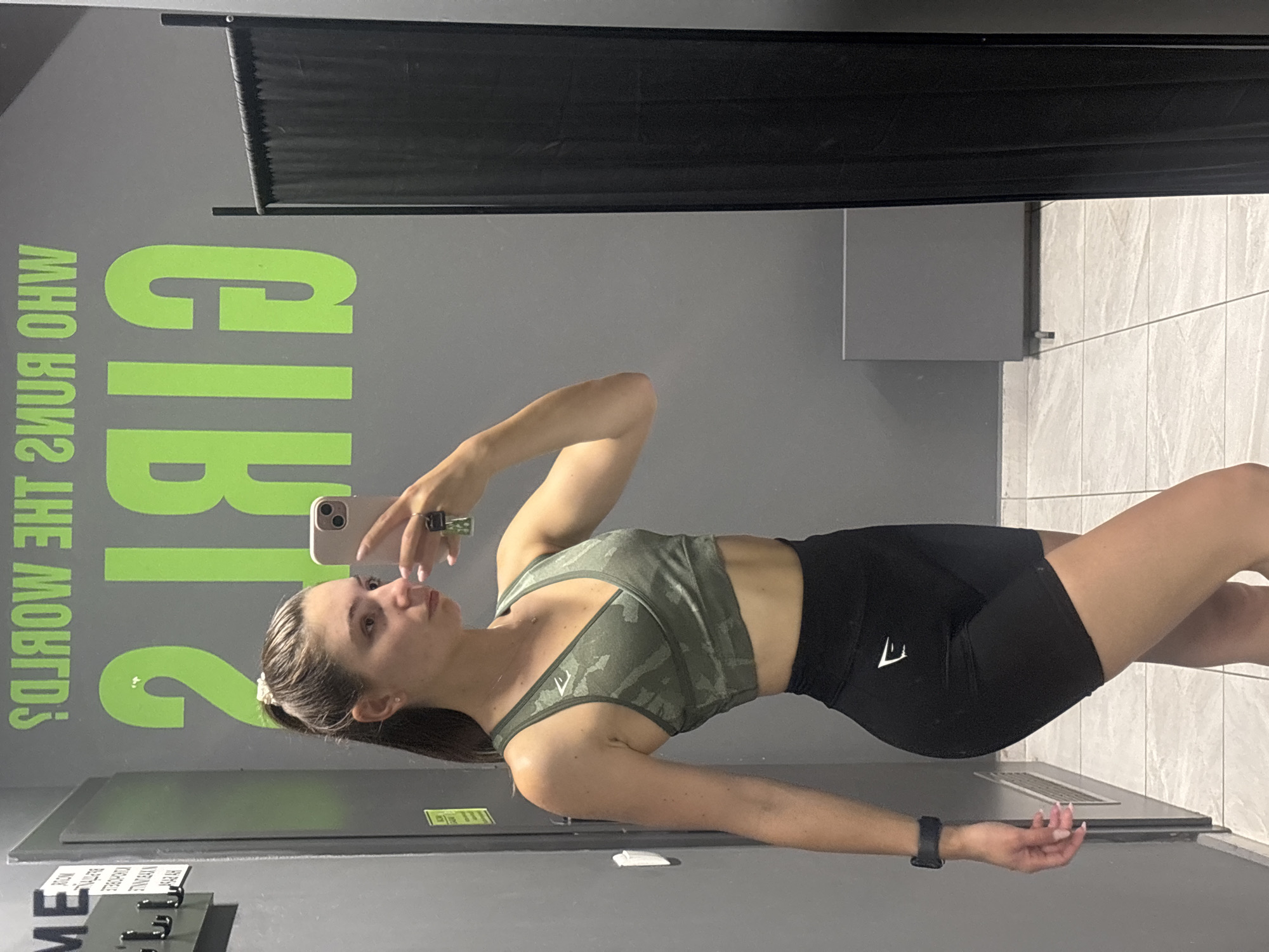 A young woman with her hair in a ponytail stands in a locker room and takes a mirror selfie. She is wearing a camouflage sports bra and black shorts. In the background, the words "GIRLS RUN THE WORLD?" are visible in large green lettering on the wall. The locker room appears to be in a gym.