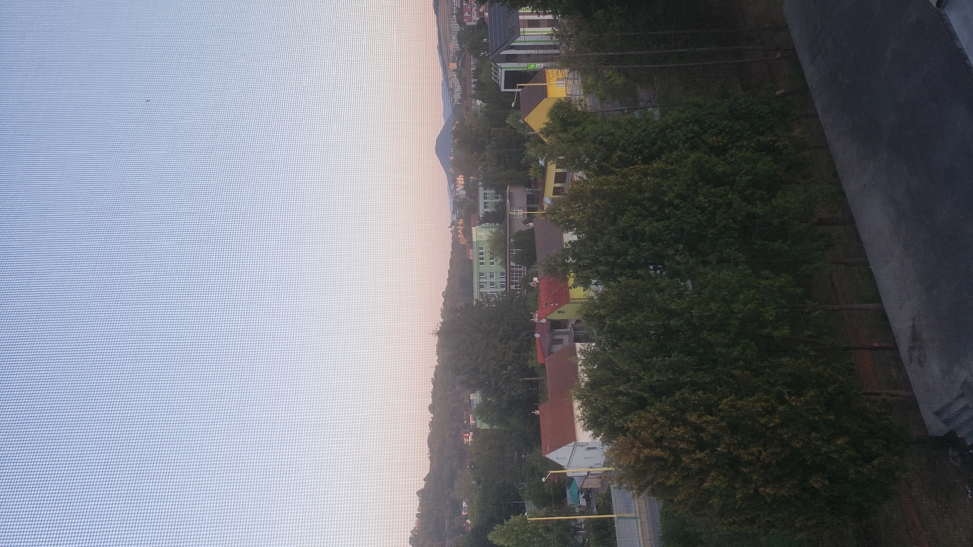 The image shows a suburban landscape at dusk, likely in Prešov, Slovakia, viewed through a window screen, indicated by the grid pattern overlaying the scene. In the foreground, a row of tall, dark green evergreen trees lines a dark, paved area, possibly a rooftop or balcony. Beyond the trees, a street is visible with a light pole and several houses, some with red tile roofs and others with yellow and white facades. Further in the background, larger buildings, including a light green structure with multiple windows and a darker building with angular architecture and colored panels, are visible. Rolling hills covered in trees form the backdrop, and a distant mountain with a rounded peak is visible on the horizon. The sky transitions from a pale pinkish hue near the horizon to a light blue overhead, suggesting the sun has recently set. The overall impression is a quiet residential area in the evening.