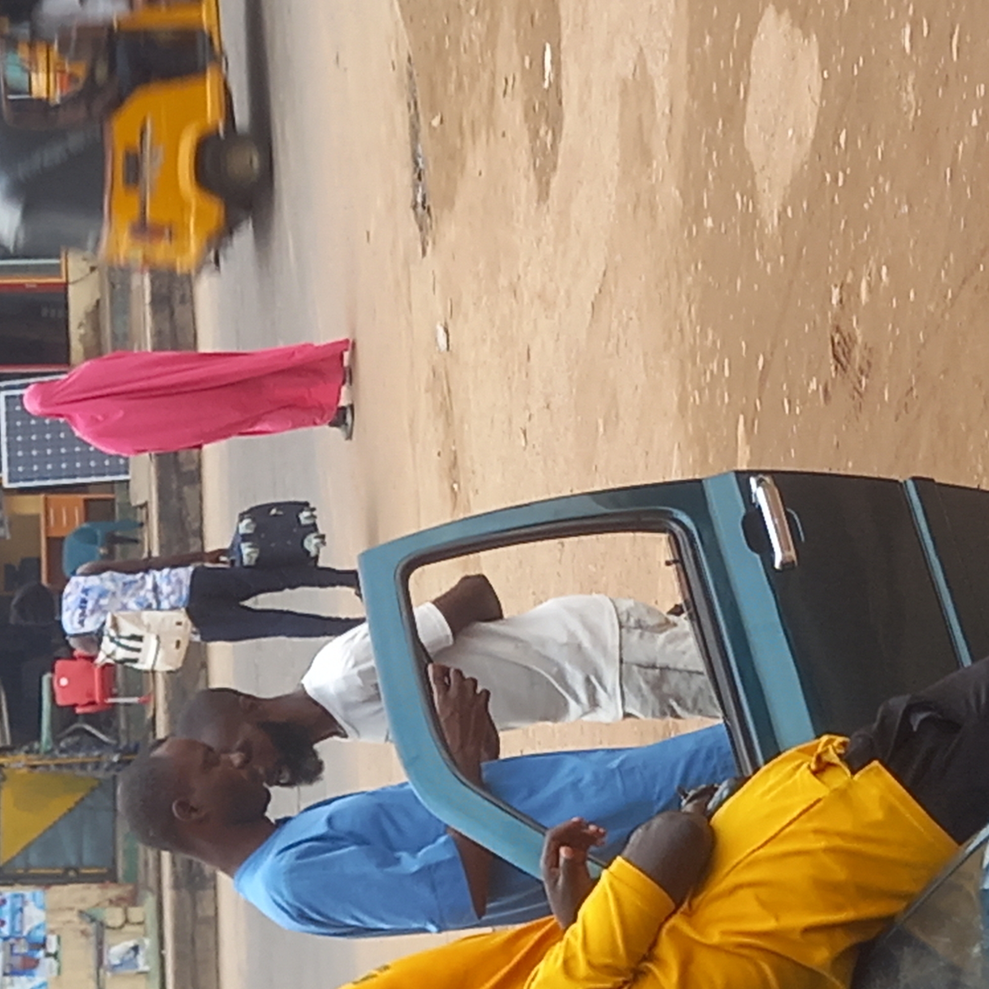 The scene captures a street in Dadi Riba, Nigeria. A partially open green car door dominates the foreground. Three men are visible near the car. One man, wearing a blue shirt, is near the car. Another man, wearing a white shirt and grey jeans, is standing by the open car door. A third man wearing a yellow shirt is seated in the car. In the background, a woman in a pink robe walks on the road. A yellow tricycle is parked on the roadside. Other people are walking in the background as well. Several buildings are visible.