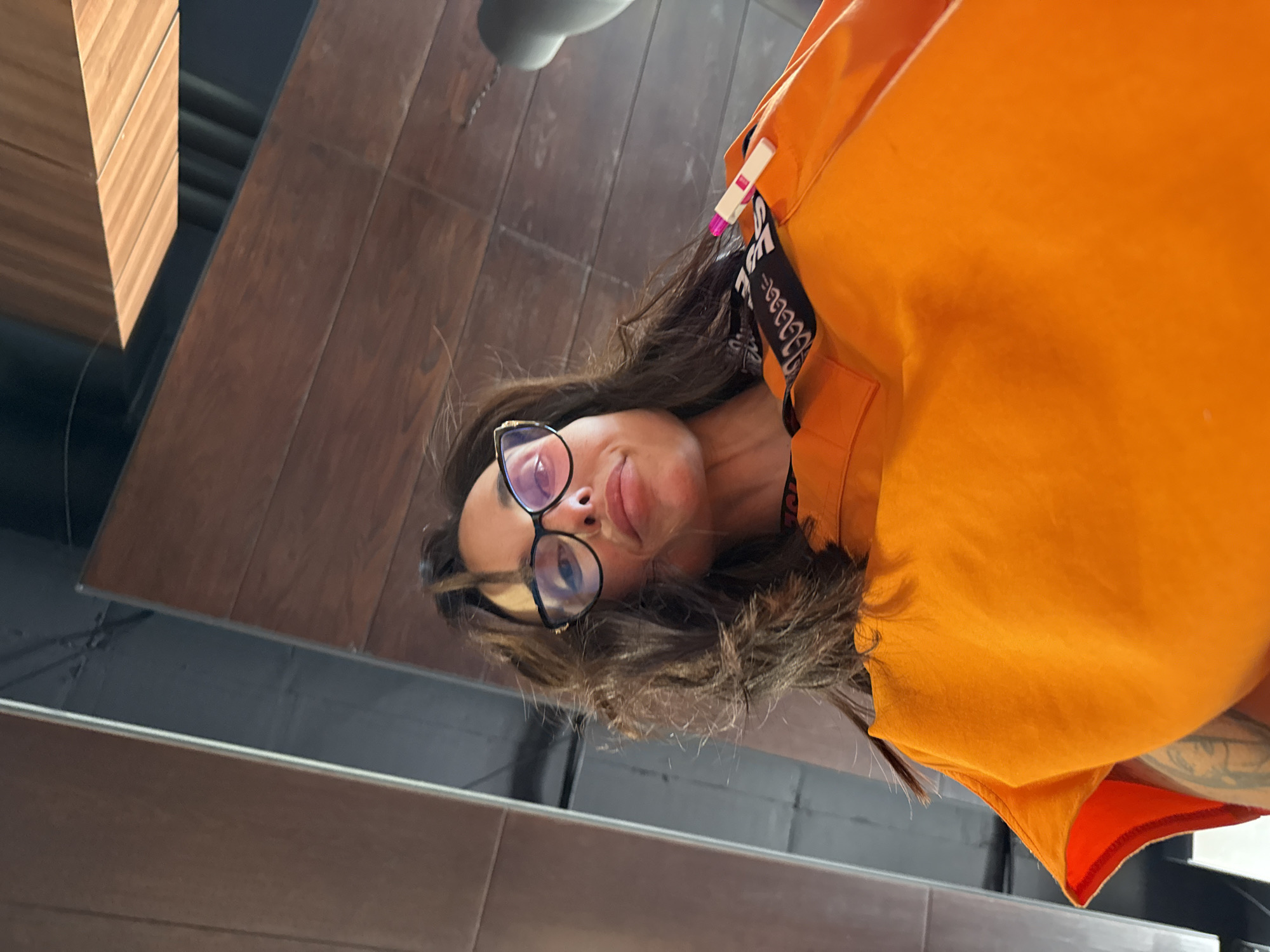 This is a close-up, low-angle selfie of a young woman with long, dark hair and glasses. She is wearing a bright orange top. Her head is tilted, and she is looking at the camera with a playful expression. There is a black strap with white text visible around her neck, possibly from a bag or accessory. A small pink and white object, resembling a clothespin or a charm, is attached to her hair. The background appears to be a reflective surface, likely a mirror, showing a wooden floor and a wooden structure on the left. The overall lighting suggests it might be daytime, possibly indoors given the reflections. The setting appears to be modern and possibly a public space, suggested by the wooden flooring and structure. The image is slightly distorted due to the low angle and the reflective surface.