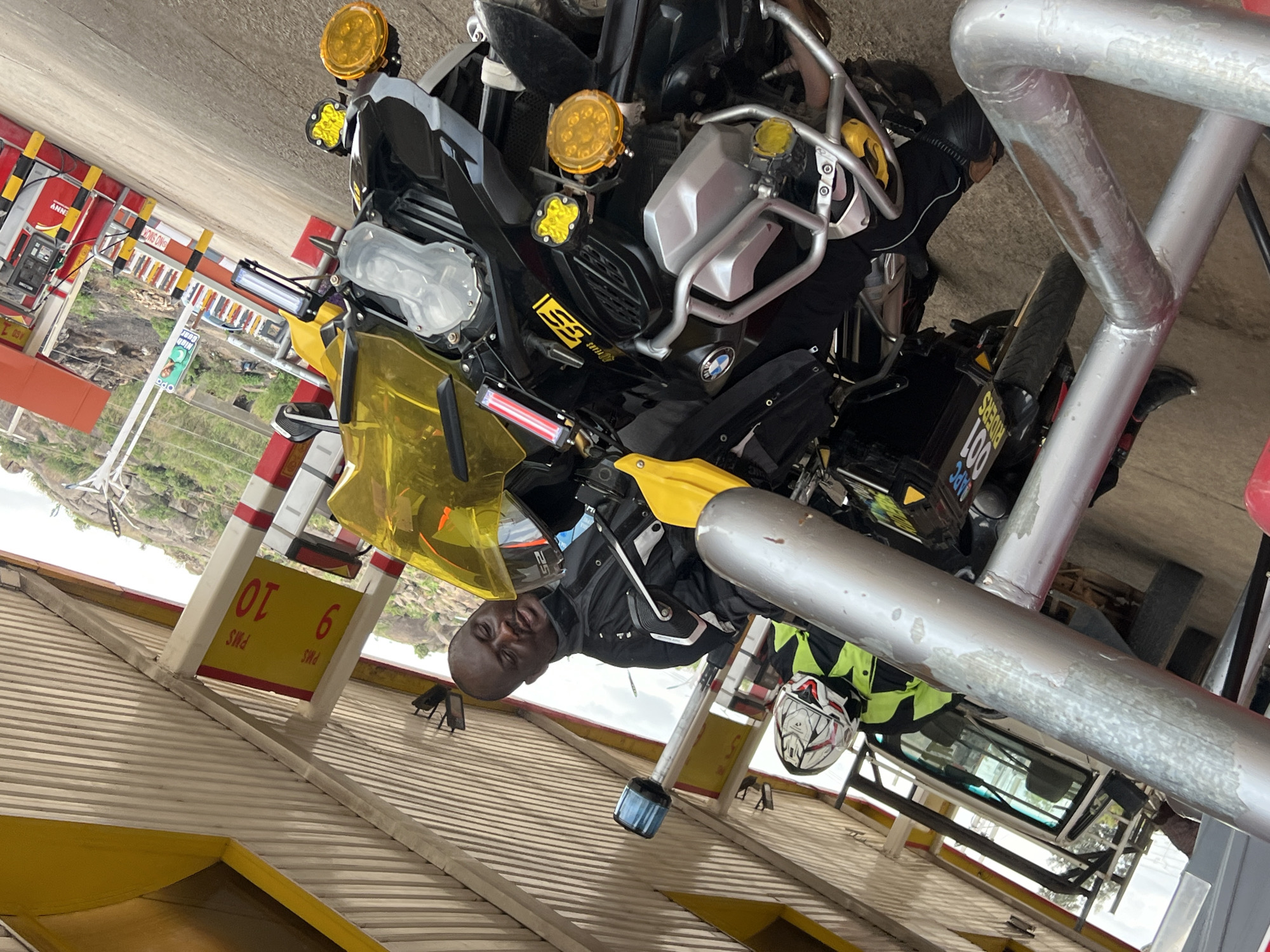 The image captures a man and a BMW motorcycle at a toll booth in Gura Top, Nigeria. The man is bald and wearing a black jacket. The motorcycle is black and yellow, with additional yellow auxiliary lights. A white helmet rests near the motorcycle. The toll booth displays "PMS 9" and "PMS 10" on its side. The environment consists of buildings, rocky hills, and foliage. A white vehicle is parked in the background.