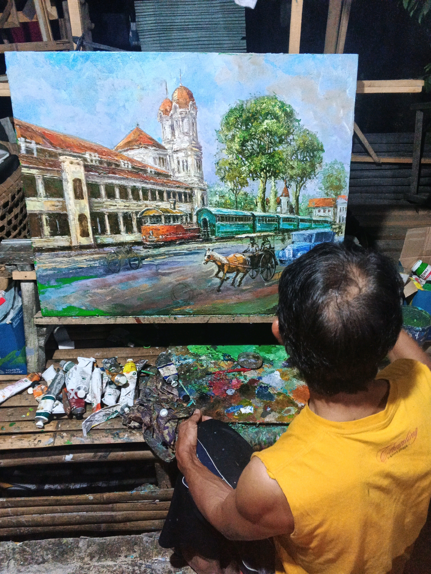 In Ungaran, Indonesia, an artist, seen from behind, is working on an oil painting of a colonial-era street scene. The painting depicts a large, ornate building with a domed tower, a tram, a horse-drawn carriage, and other vehicles. The artist, wearing a yellow t-shirt, is seated in front of the painting on a wooden surface cluttered with tubes of paint, brushes, and a palette. The surrounding environment appears to be an outdoor workshop or studio, with wooden structures and foliage visible in the background.