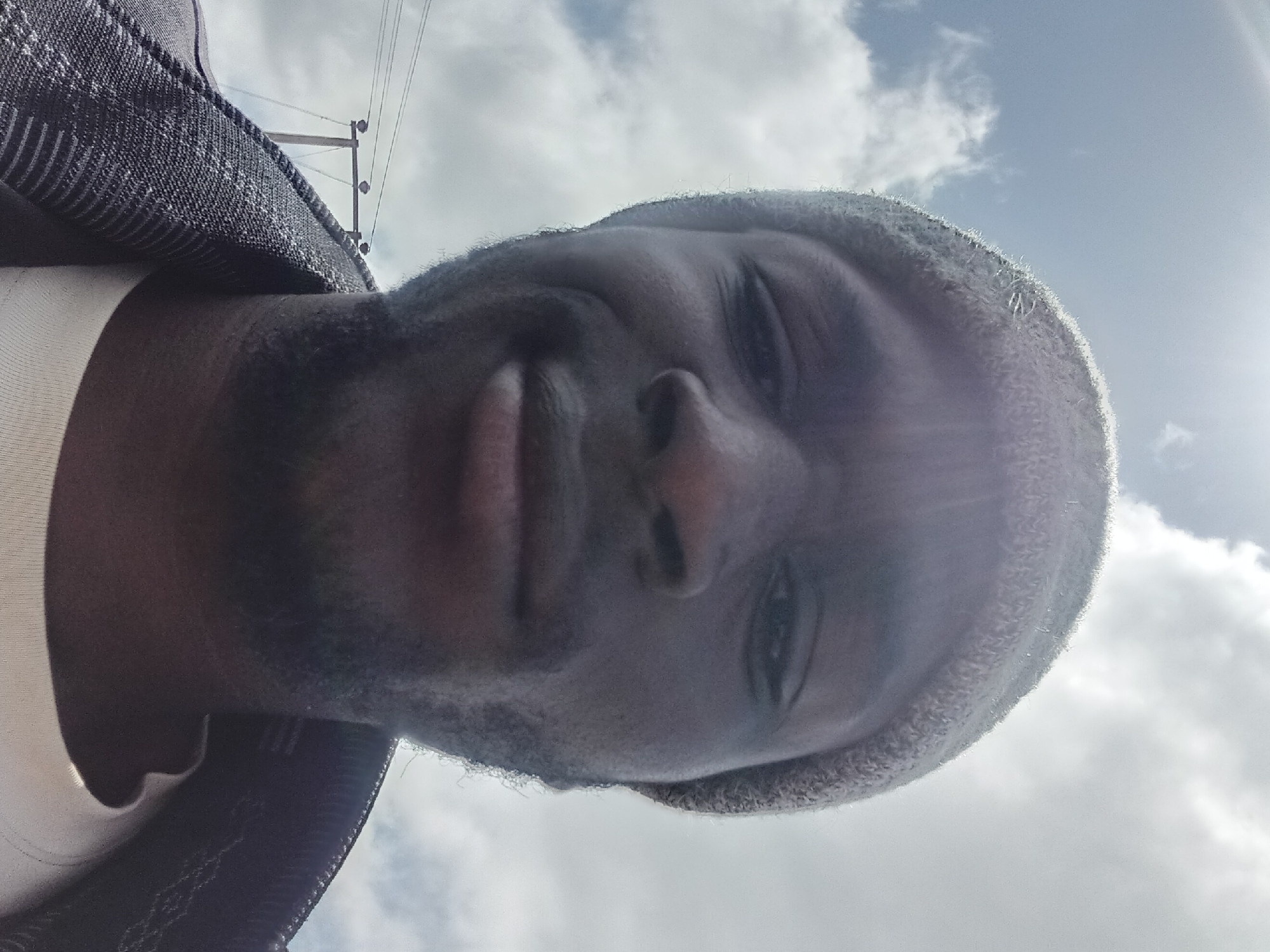 The image is a close-up, low-angle selfie of a Black man wearing a grey knit beanie and a dark jacket with a textured pattern over a white t-shirt. He has short facial hair and is looking slightly off-camera with a neutral expression. The background consists of a bright sky with scattered clouds. In the upper left portion of the frame, a utility pole with wires is visible. The scene appears to be outdoors, possibly in an urban or semi-urban setting within Bukuru, Nigeria. The lighting suggests it is daytime.