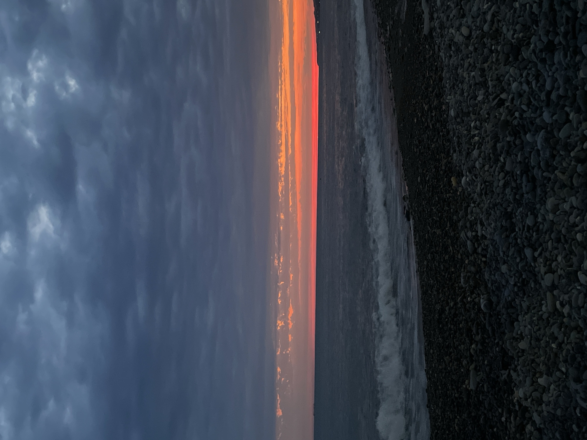 This image depicts a twilight scene on a rocky beach. The sky is a dramatic gradient of dark, cloudy gray at the top, transitioning to bands of orange and red near the horizon. The ocean is a deep, dark blue with white waves crashing onto the shore. The foreground is dominated by a dense scattering of dark, smooth stones and pebbles that make up the beach. The image is devoid of any people or man-made structures, focusing solely on the natural landscape.