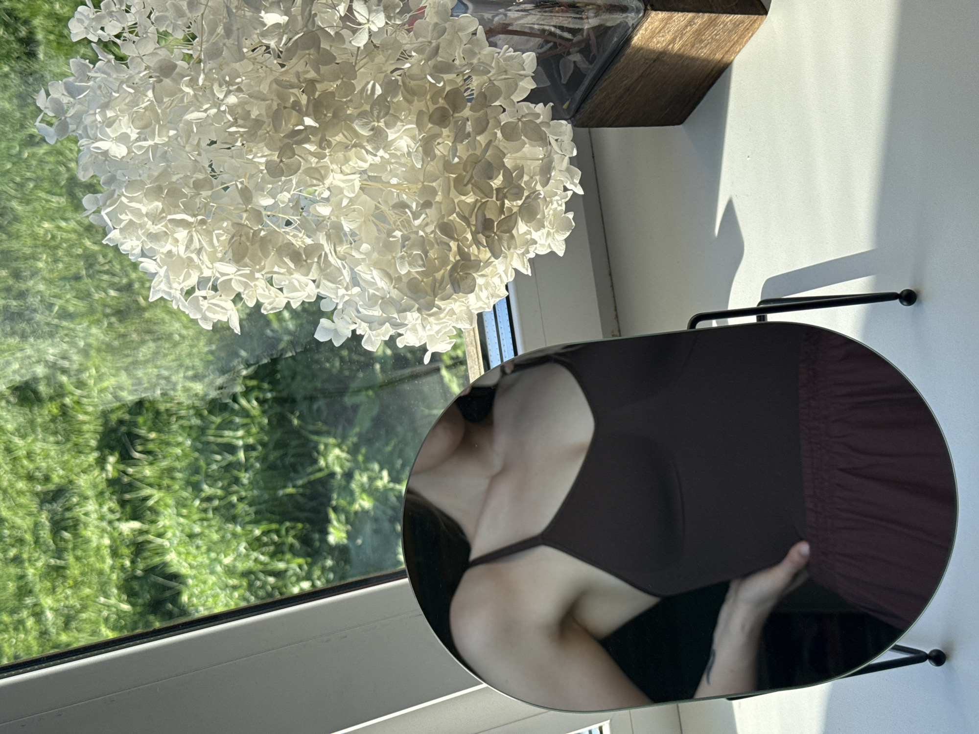 The image depicts an indoor scene, possibly in a home, featuring a round mirror reflecting a partial view of a person. The person appears to be wearing a dark garment, likely a dress or top with a thin strap, and is holding or adjusting the clothing. The background of the reflection shows a window with an exterior view of greenery.
The mirror is positioned on a white surface, possibly a windowsill or table, next to a bouquet of white flowers. Above the flowers is a wooden object. Outside the window, green trees and grass are visible.
There is no explicit evidence to confirm the location is Chaikovsky, Russia.