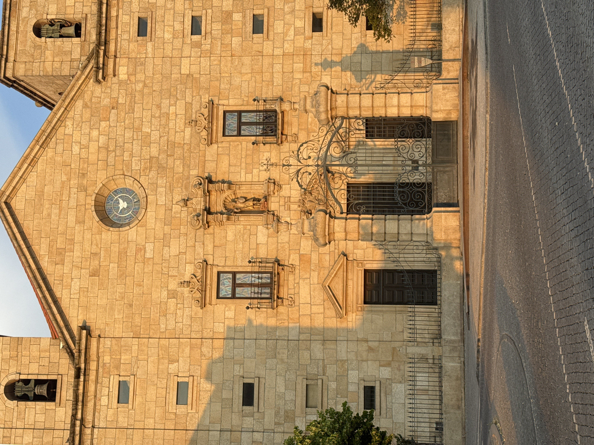 The image shows a close-up, eye-level view of the facade of a stone church, illuminated by the warm, golden light of what appears to be late afternoon or early morning sun. The architecture is detailed, with many small, rectangular windows, some with decorative stained glass, and larger arched openings. The main entrance is a tall, dark wooden door framed by an elaborate stone archway and surmounted by a wrought-iron gate. Above the entrance, a niche contains a statue of a religious figure, and a circular stained-glass window is positioned higher on the facade. Two bell towers, one on each side of the main structure, are visible in the upper corners, each with an arched opening housing a bell. The church is built with rough-hewn, light-colored stone blocks. In front of the building, a dark wrought-iron fence runs horizontally, separating the church from a cobblestone street. A small tree with green foliage is visible to the left, and the shadow of another tree falls on the right side of the facade. The cobblestone street in the foreground displays a pattern of tightly packed stones and faint white lines, suggesting a road intersection. The overall impression is one of historical architecture bathed in natural light. This is the Church of Saint Mary of the Snows (Igreja de Santa Maria das Neves) located in the city of Castelo Branco, Portugal.