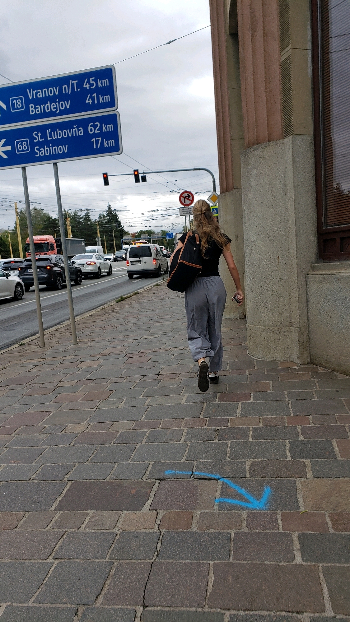 The image captures a street scene in Prešov, Slovakia, looking eastward along a cobblestone sidewalk. A young woman with blonde hair tied in a ponytail is walking away from the camera, carrying a dark bag over her left shoulder. She is wearing a black t-shirt and light gray, loose-fitting pants. Her right hand is extended slightly as she walks. On the sidewalk in the foreground, a bright blue arrow is spray-painted, pointing to the lower right.
Beyond the sidewalk, a two-lane road carries moderate traffic, including several cars and a white van. On the left side of the road, a large blue road sign with white lettering indicates directions to "Vranov n/T. 45 km," "Bardejov 41 km," "St. Ľubovnía 62 km," and "Sabinov 17 km." Traffic lights and electrical wires are visible above the road. The architecture on the right side of the image features a building with ribbed columns and a window. The sky is overcast, suggesting a cloudy day.