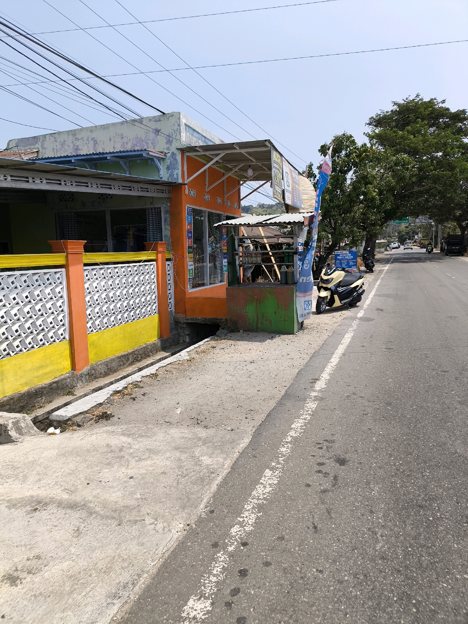 The image depicts a street scene in Indonesia, likely a commercial area. A building with an orange facade and a white awning is on the left, adjacent to a yellow and orange fence with decorative white concrete blocks. A small kiosk with a green base stands next to the building, with items displayed on shelves. A beige scooter is parked in front of the kiosk. The street is paved with asphalt and has a white line marking. Trees line the right side of the street, and in the background, there are other buildings and vehicles. The sky is clear and blue.